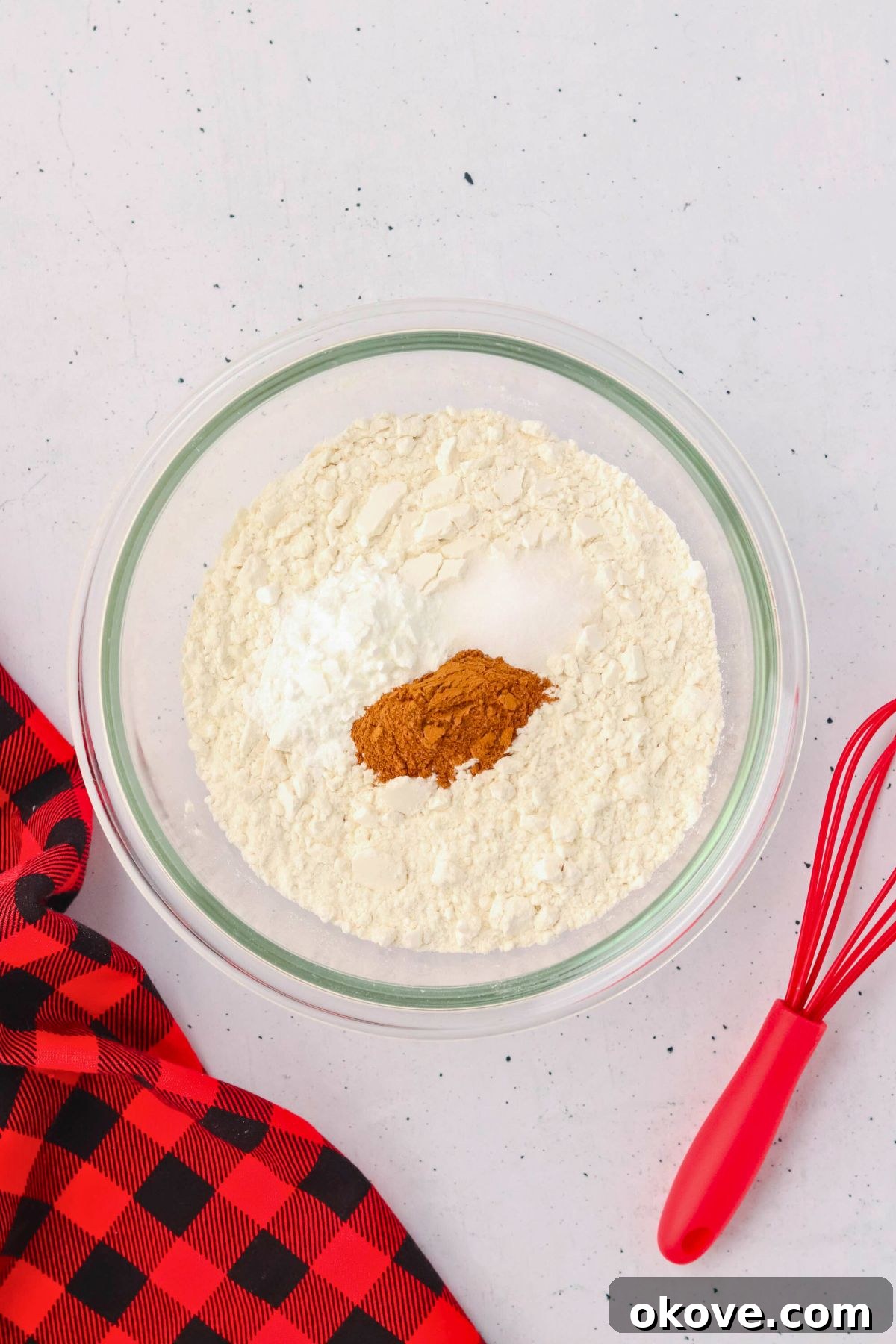 Dry ingredients including flour, baking powder, cinnamon, and salt being mixed together in a clear glass bowl using a whisk.