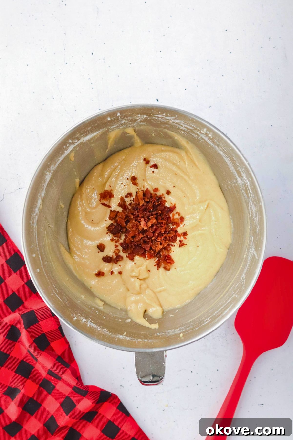 Cupcake batter being gently folded with crispy bacon bits using a vibrant red spatula in a glass bowl.