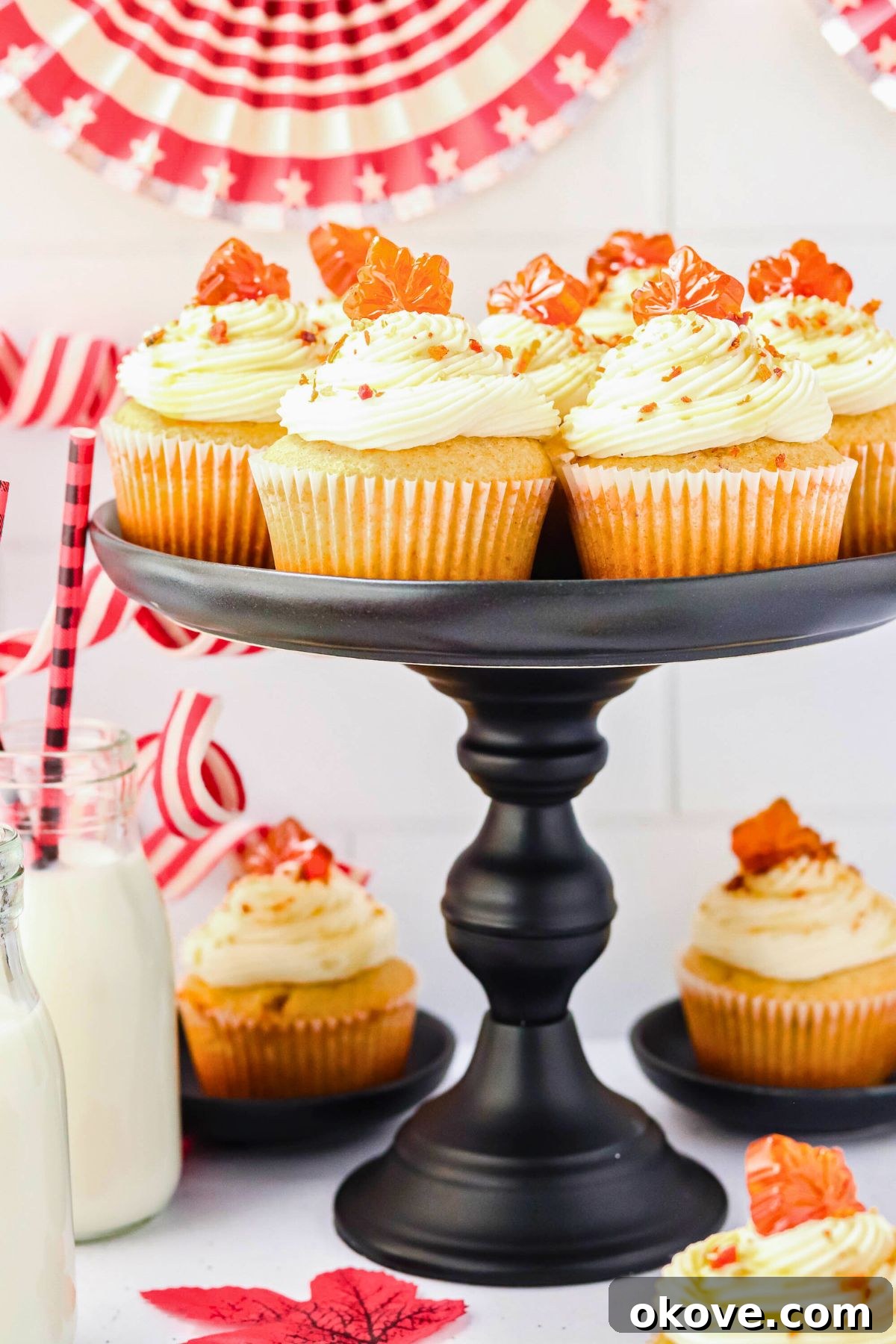 Beautifully frosted maple bacon cupcakes displayed on a black cake stand, with a glass of milk and more cupcakes on a white marble table in the background.