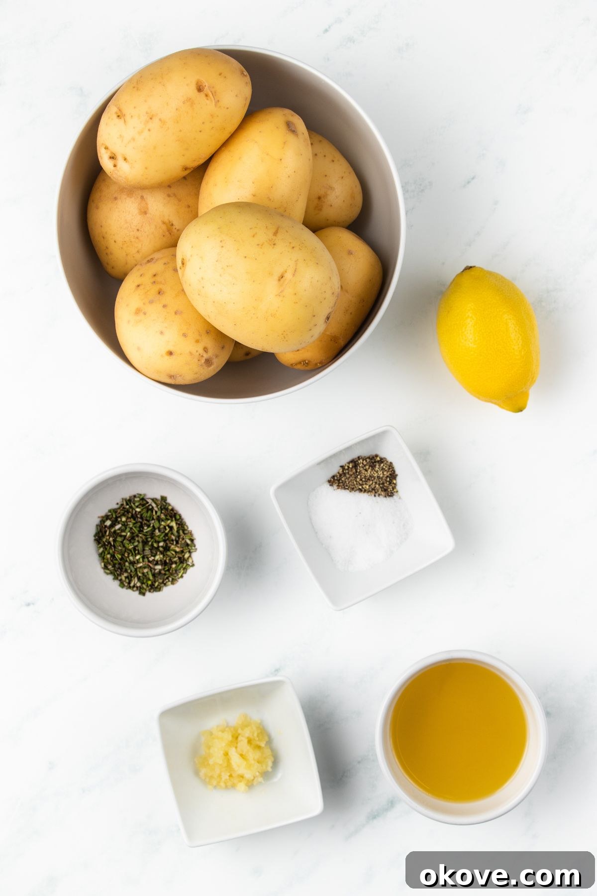 The Ultimate Roasted Spuds 3 Ingredients for roasted potatoes laid out on a white marble surface, including whole potatoes, olive oil, spices, rosemary, garlic, and lemon.