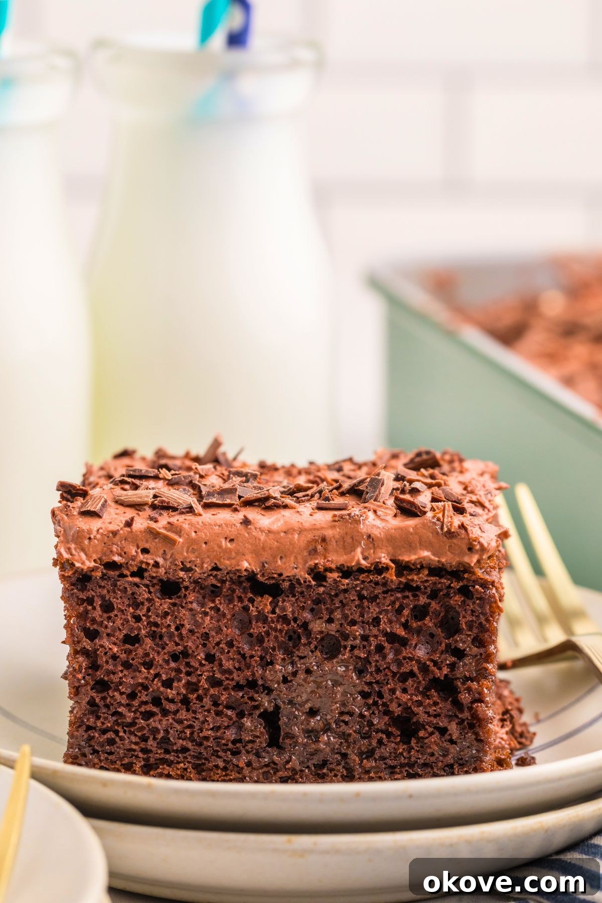 A slice of luscious chocolate poke cake on a white plate, with decorative milk bottles in the background. The cake features visible layers of moist chocolate cake, a creamy white topping, and chocolate shavings.