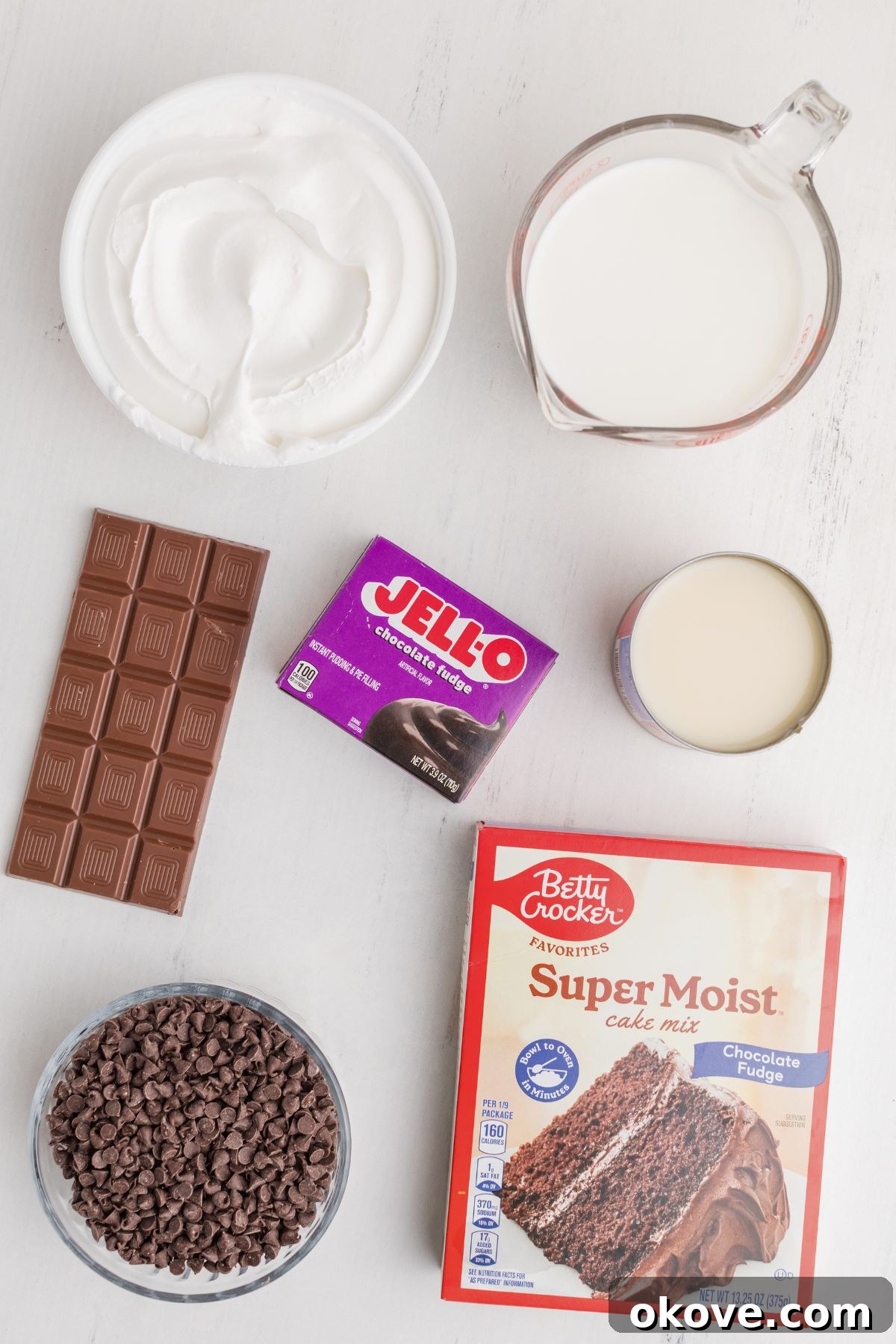 A collection of ingredients laid out on a white marble table, including a box of chocolate cake mix, canned sweetened condensed milk, chocolate chips, whipped topping, instant pudding mix, and a chocolate bar, all ready for baking.