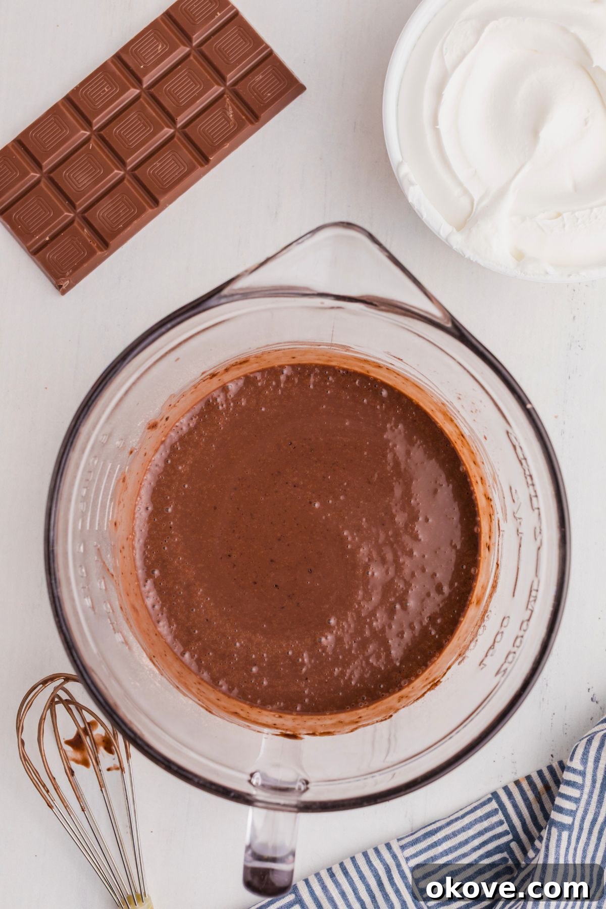 A clear glass bowl filled with rich, dark chocolate cake batter, resting on a pristine white marble countertop, ready for baking.