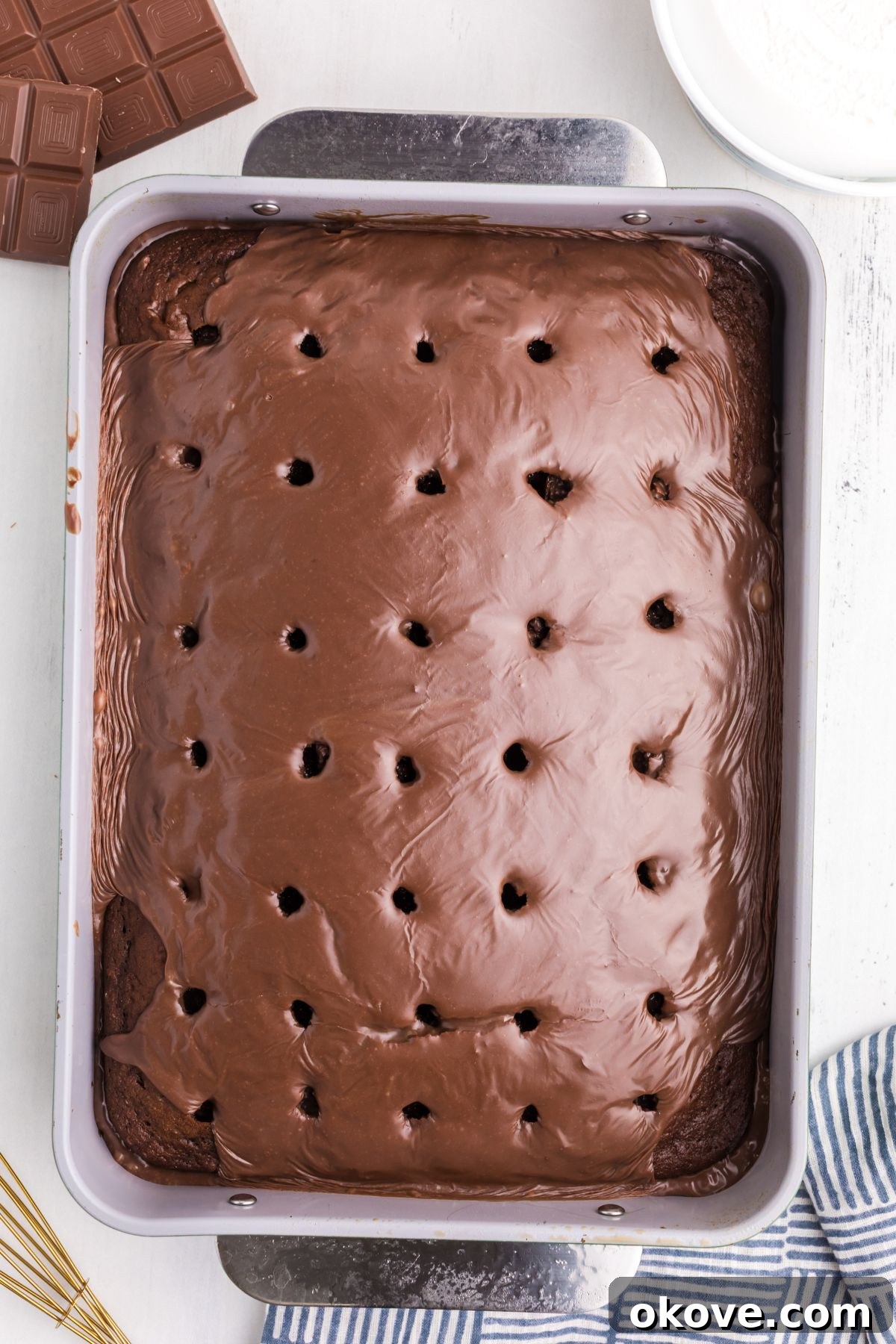 A chocolate cake, freshly poked with holes, is being generously drizzled with a smooth mixture of melted chocolate and sweetened condensed milk, sinking into each opening.