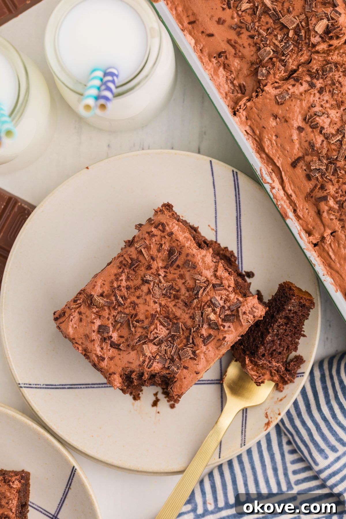 A beautifully presented slice of chocolate poke cake on a crisp white plate, positioned in front of charming glasses of milk adorned with blue straws, ready to be enjoyed.