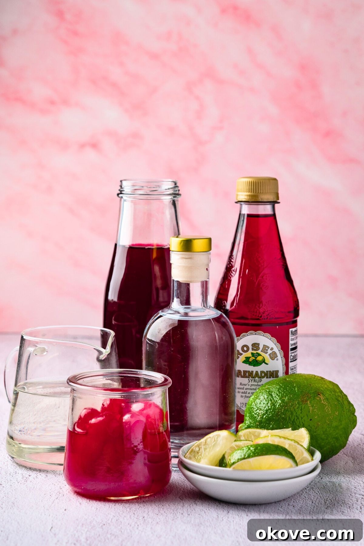 Pomegranate Martini Bliss 3 A curated selection of ingredients laid out on a clean marble table, including a bottle of premium vodka, fresh pomegranates and their juice, bright green limes, a bottle of grenadine, and a small jug of simple syrup, all artfully arranged for making a pomegranate martini.