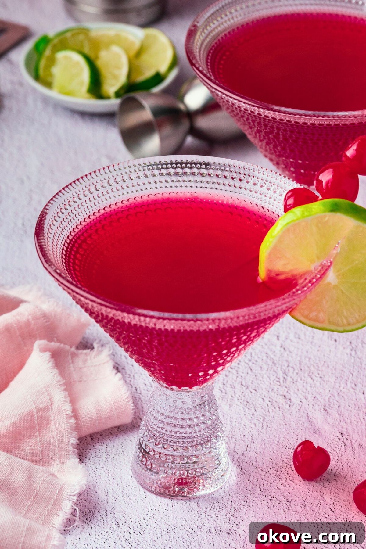 Pomegranate Martini Bliss 6 Close-up shot of a perfectly crafted pomegranate martini, vibrant red in a martini glass, with a lime wedge and a maraschino cherry on the rim, showcasing its elegant presentation.