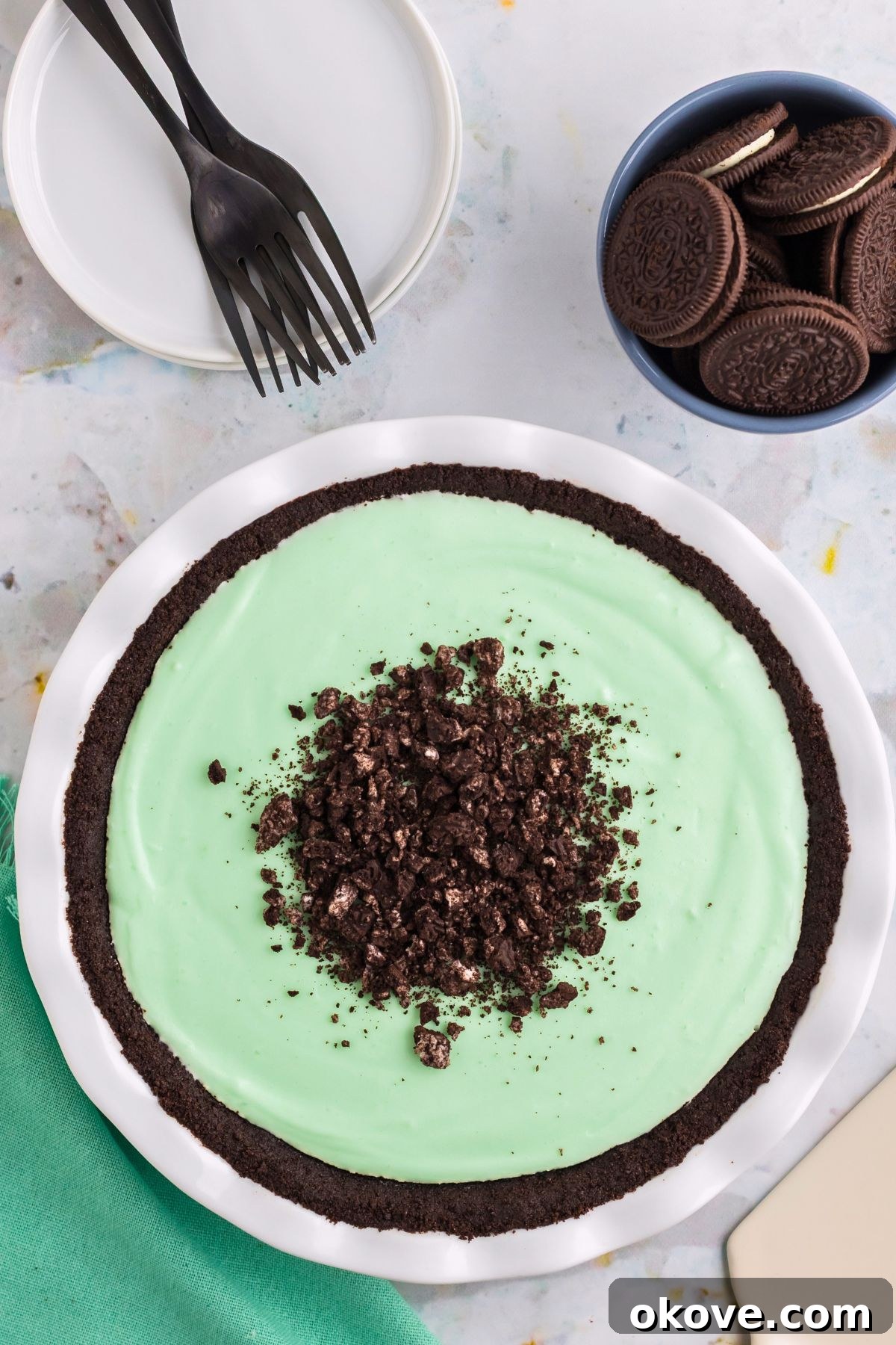 Minty Green Dream Pie 4 A luscious Grasshopper Pie, still in its white ceramic pie dish, adorned with a scattering of chocolate sandwich cookie crumbles on top, resting on a rustic wooden table.
