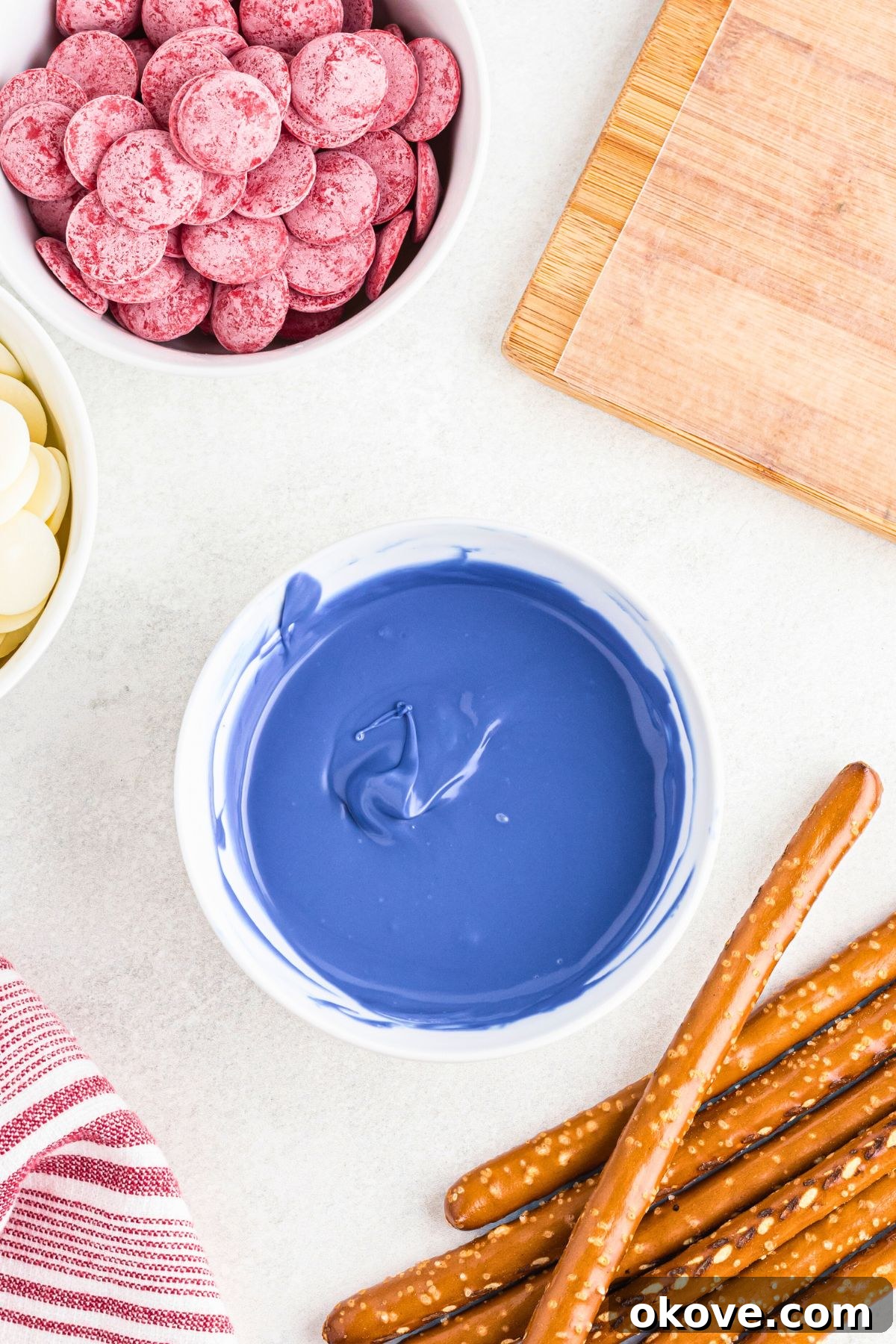 Bright blue melting chocolate melted in a small white bowl in front of pretzel rods, before being dipped.