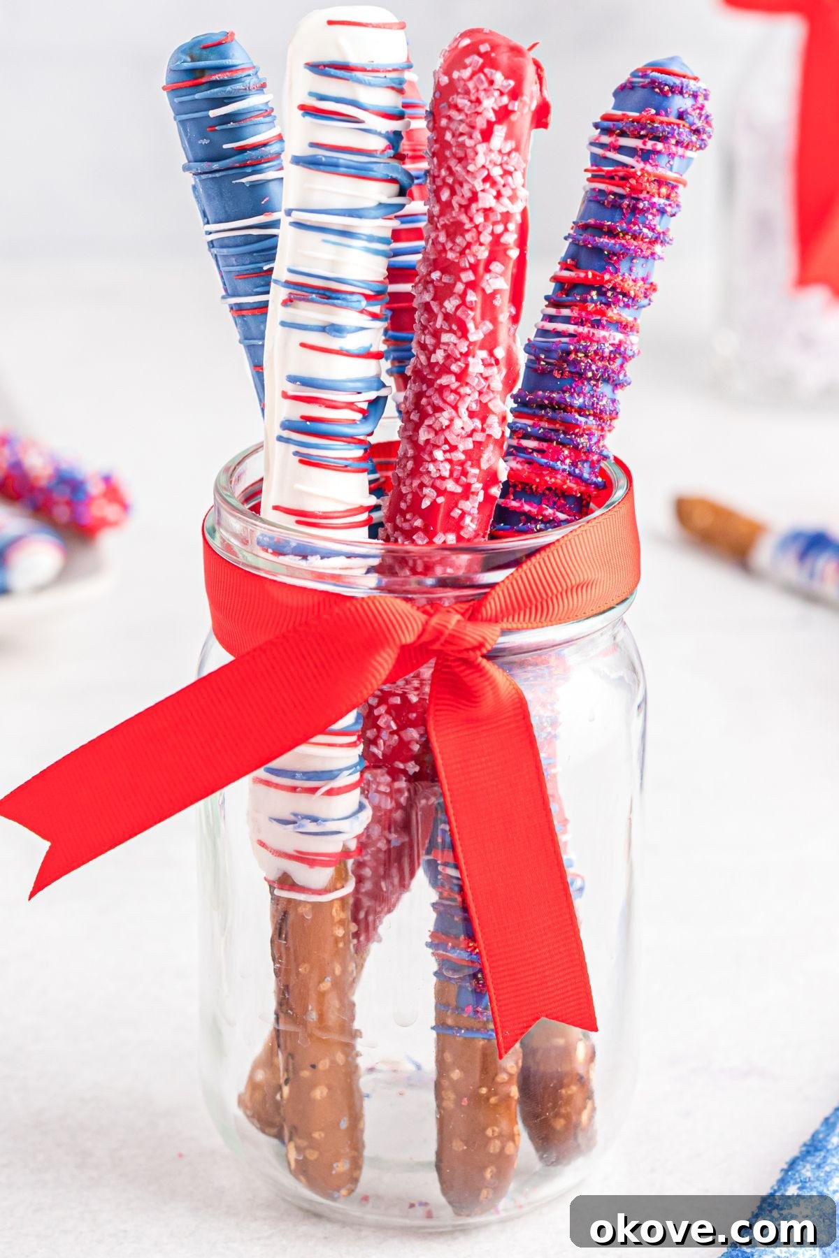 Red, white, and blue pretzels dipped in coating and sprinkles in a clear glass jar with a red ribbon, ready for serving.