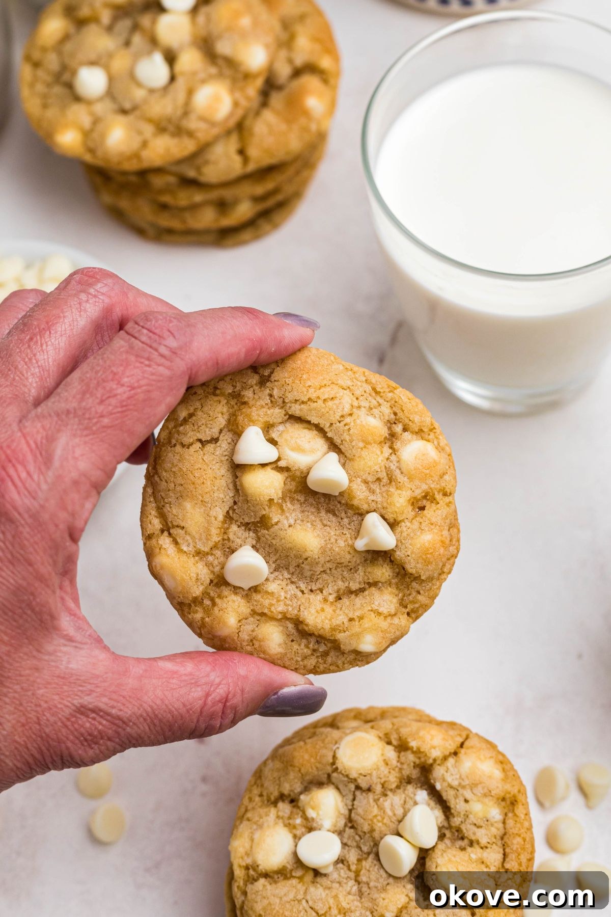 A delicious white chocolate chip cookie being held in a hand, perfectly framed against a refreshing glass of milk.