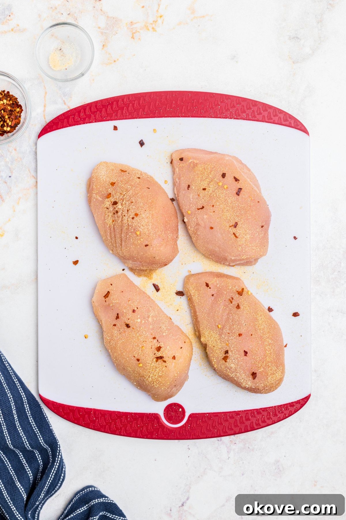 Say Yes Chicken 4 Uncooked chicken breasts being seasoned with spices on a clean white cutting board, ready for searing.
