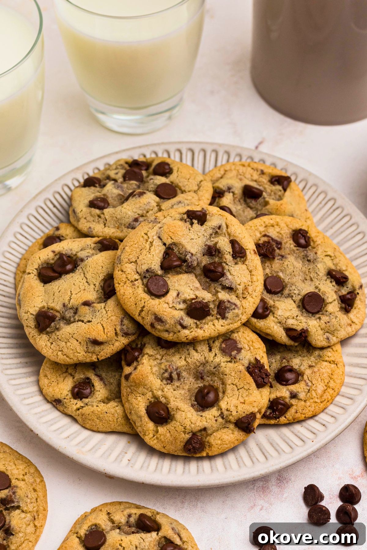 Warm Chocolate Chip Indulgence 2 Golden brown chocolate chip cookies stacked on a white plate with chocolate chips scattered on a marble surface.