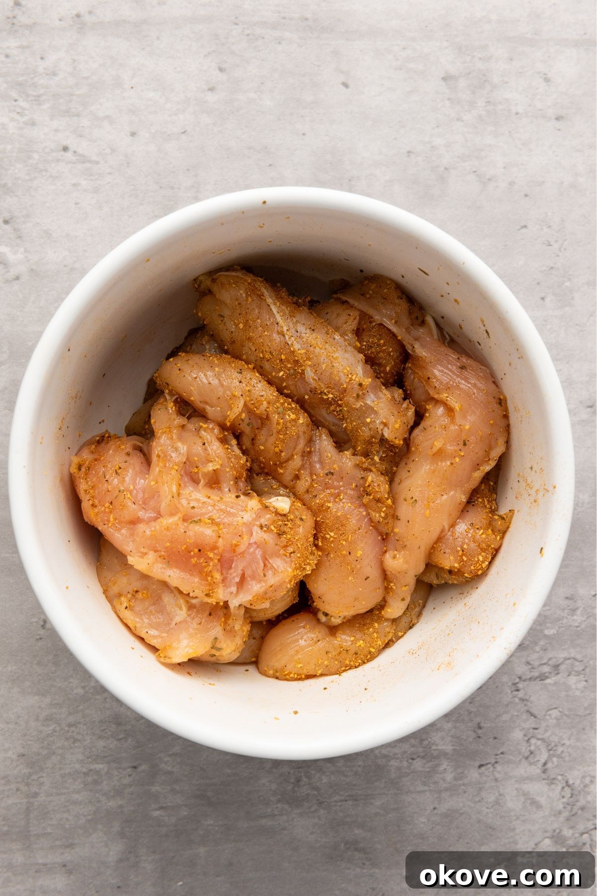 Uncooked chicken tenderloins seasoned generously with Cajun spices in a white bowl, ready for cooking.