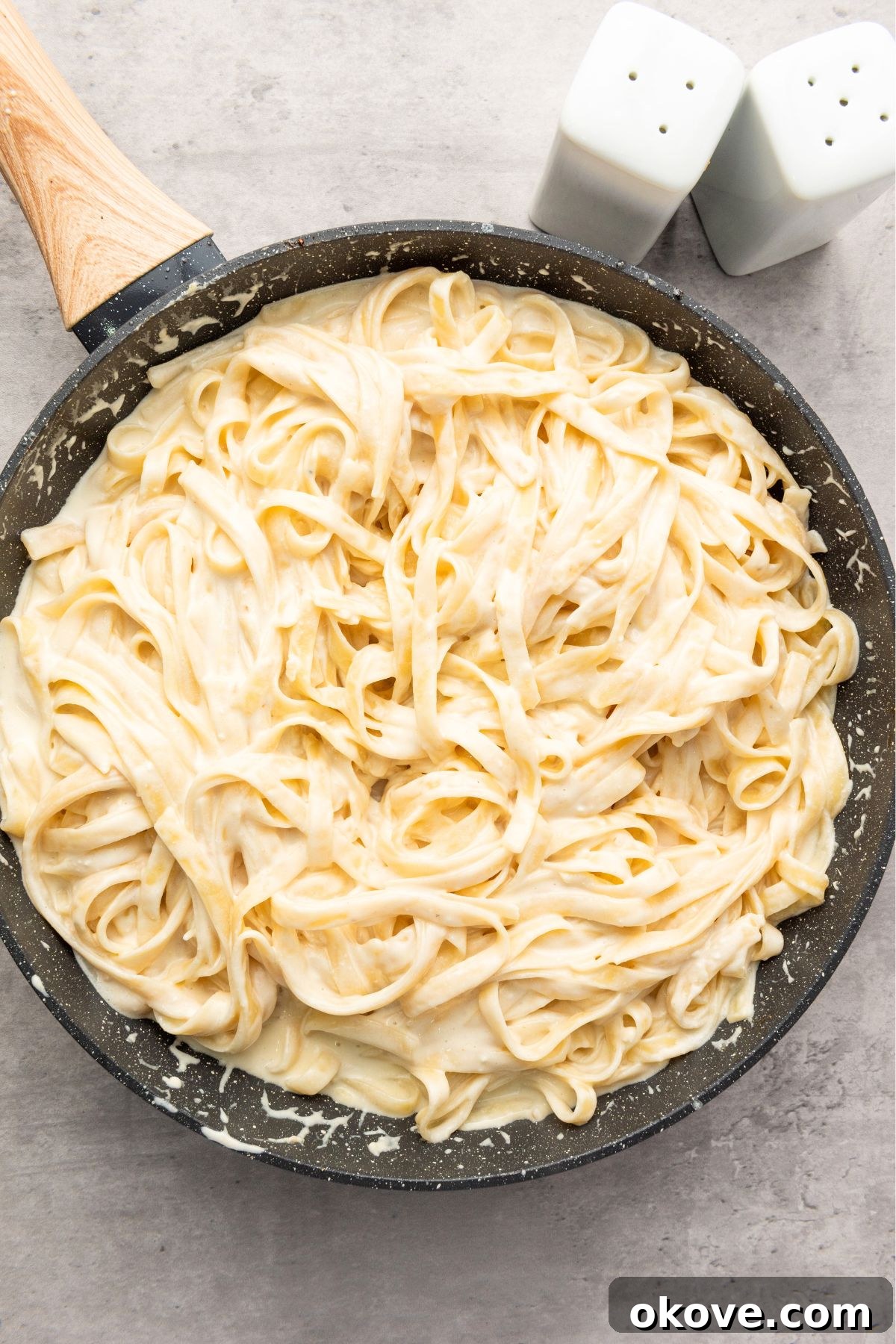 Cooked fettuccine pasta being tossed generously with creamy Alfredo sauce in a large skillet, ensuring every strand is coated.