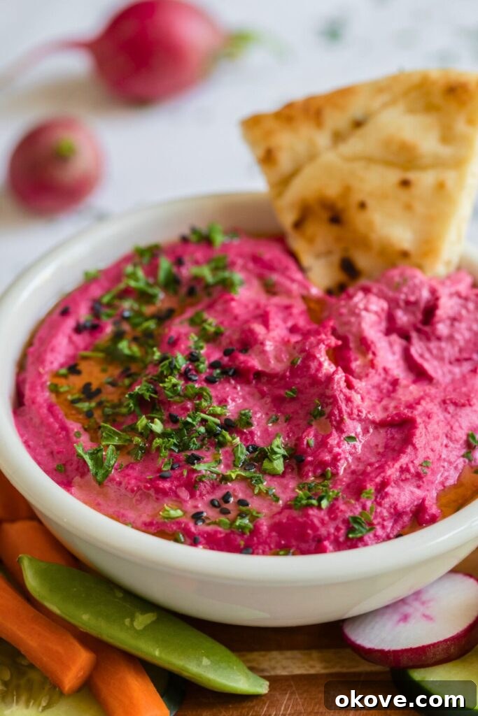 Vibrant Beet Hummus 2 Vibrant pink beet hummus in a stylish white bowl, garnished with fresh herbs and surrounded by colorful crudités like carrots, cucumbers, and radishes, ready for dipping.