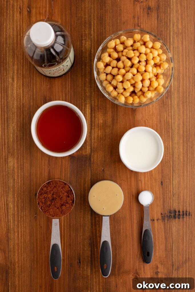 Decadent Chocolate Hummus 3 Carefully measured ingredients for chocolate hummus, including chickpeas, cocoa powder, maple syrup, tahini, and milk, arranged on a rustic wooden table.