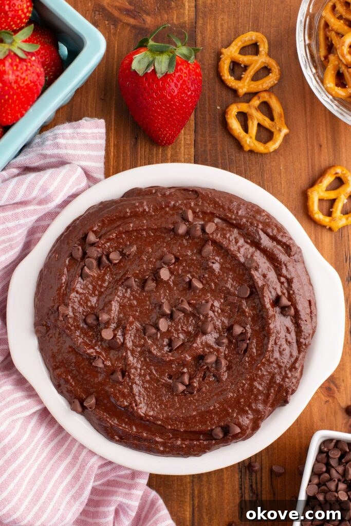 Decadent Chocolate Hummus 6 Chocolate hummus in a white serving dish, topped with mini chocolate chips, surrounded by strawberries and pretzels for dipping.