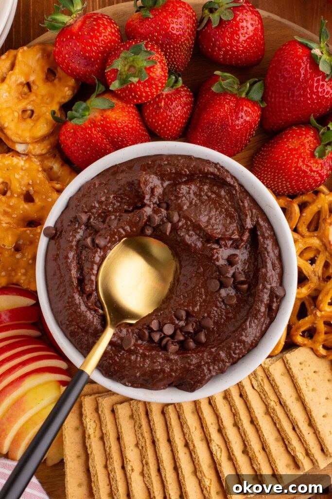 Decadent Chocolate Hummus 8 A elegant white bowl of chocolate hummus, with a golden spoon perfectly placed, surrounded by an enticing arrangement of fresh strawberries and salty pretzel sticks for dipping.
