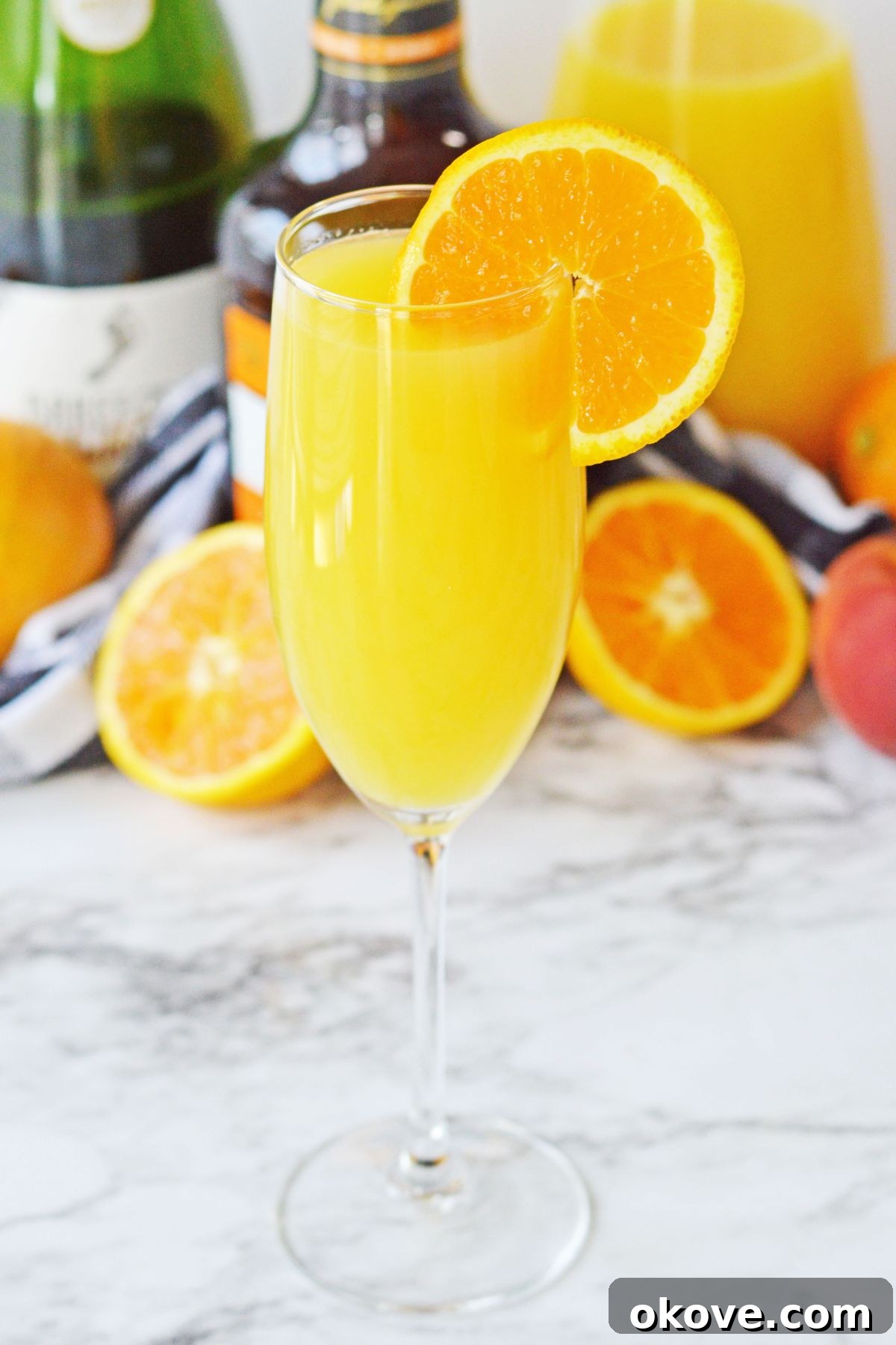 Sunrise Peach Sparkler 2 A vibrant peach mimosa, garnished with a fresh orange slice, resting on a pristine white marble table. The drink appears perfectly chilled and sparkling.