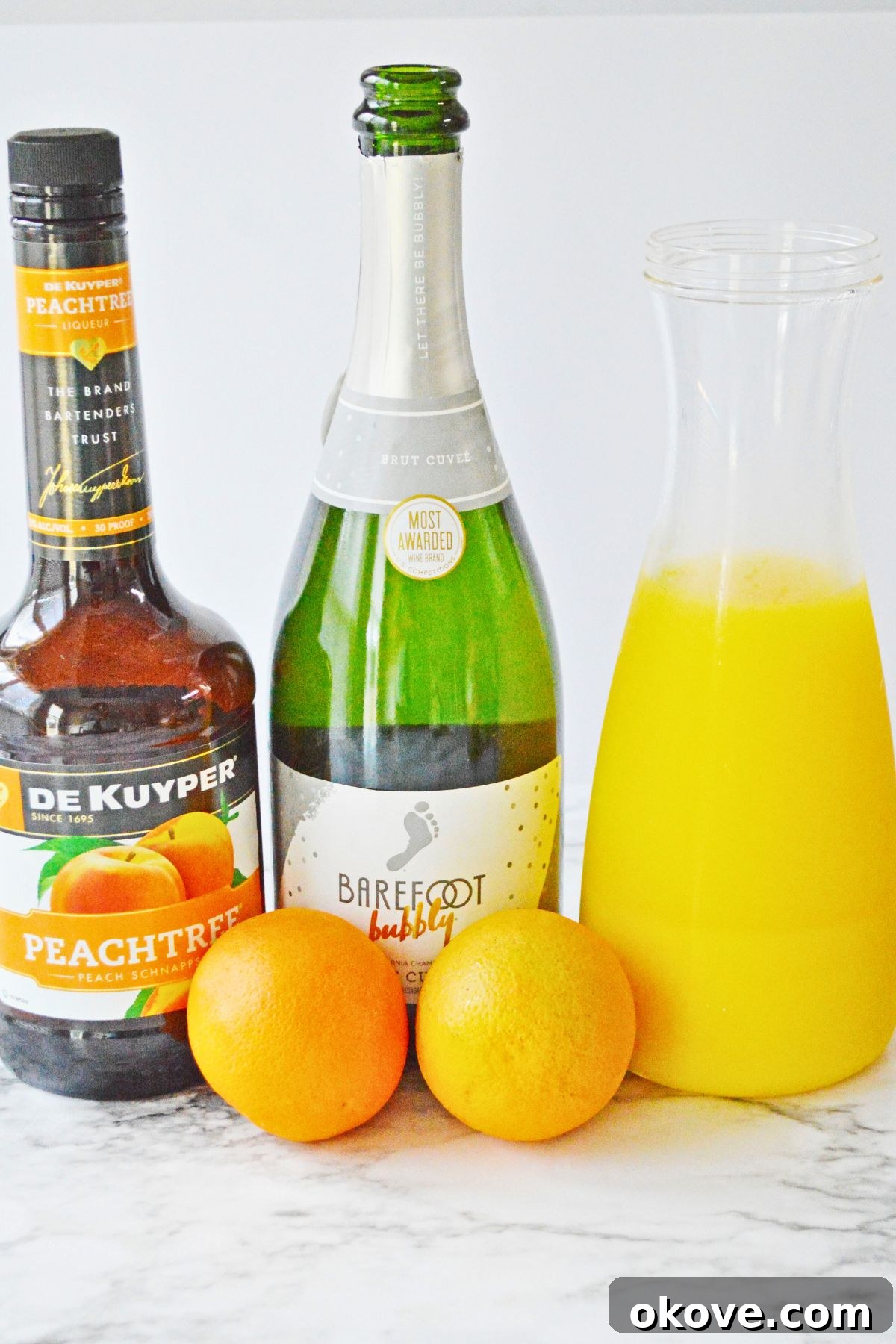 Sunrise Peach Sparkler 3 Fresh ingredients for making peach mimosas, including a bottle of peach schnapps, orange juice, and an orange for garnish, arranged on a white marble surface.