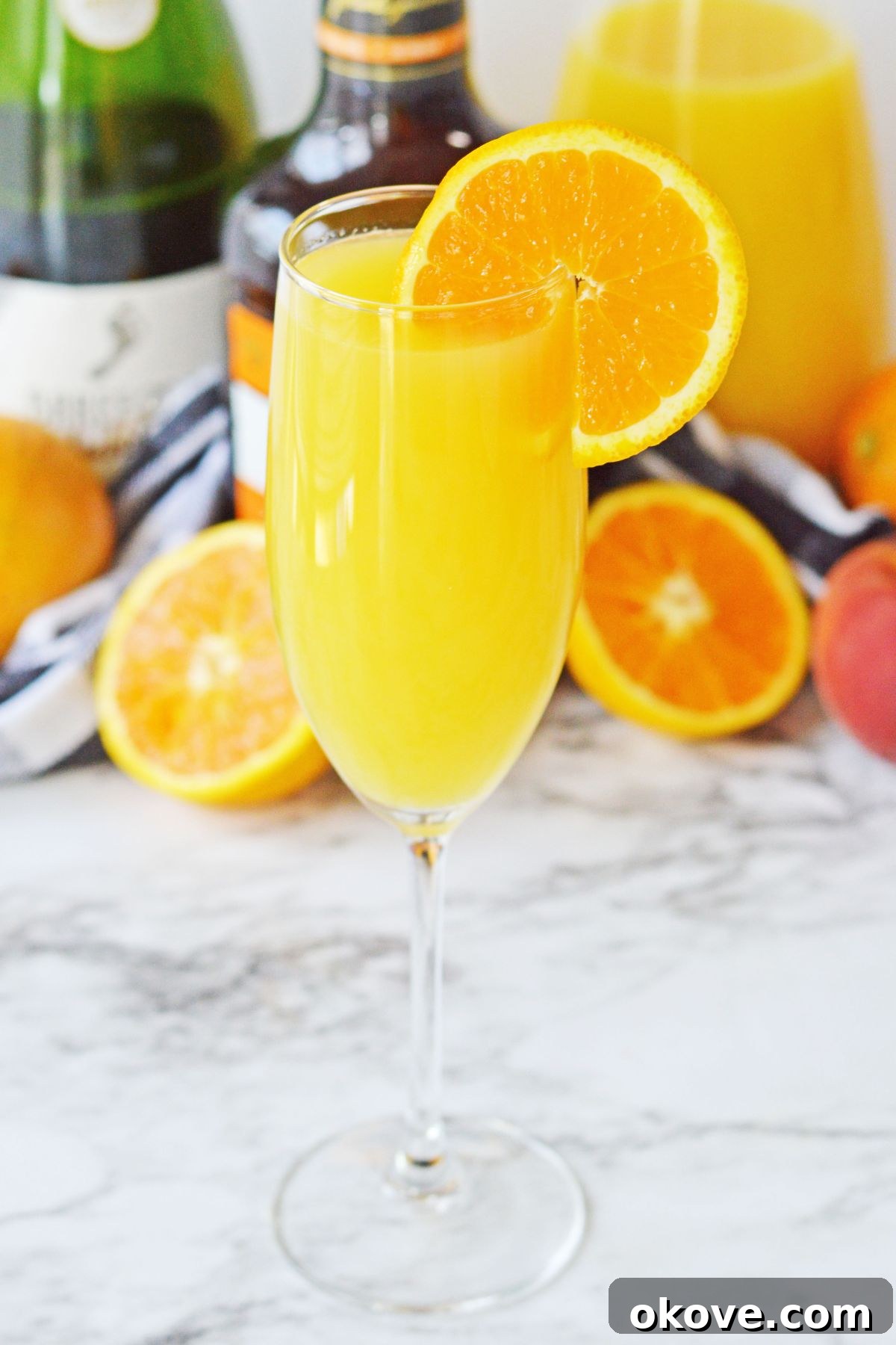 Sunrise Peach Sparkler 5 A close-up shot of a peach mimosa in a tall flute, garnished with a fresh orange slice, set against a backdrop of white marble.