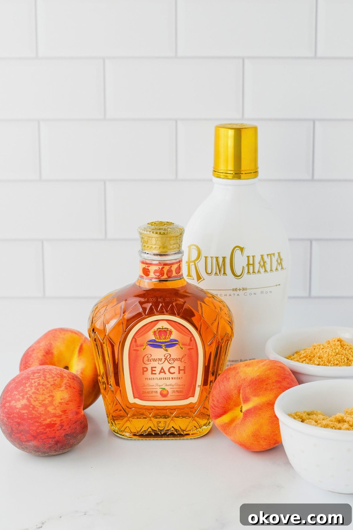 Ingredients needed to make a peach cobbler shot laid out on a clean marble table, including whiskey, rumchata, graham crackers, and brown sugar.