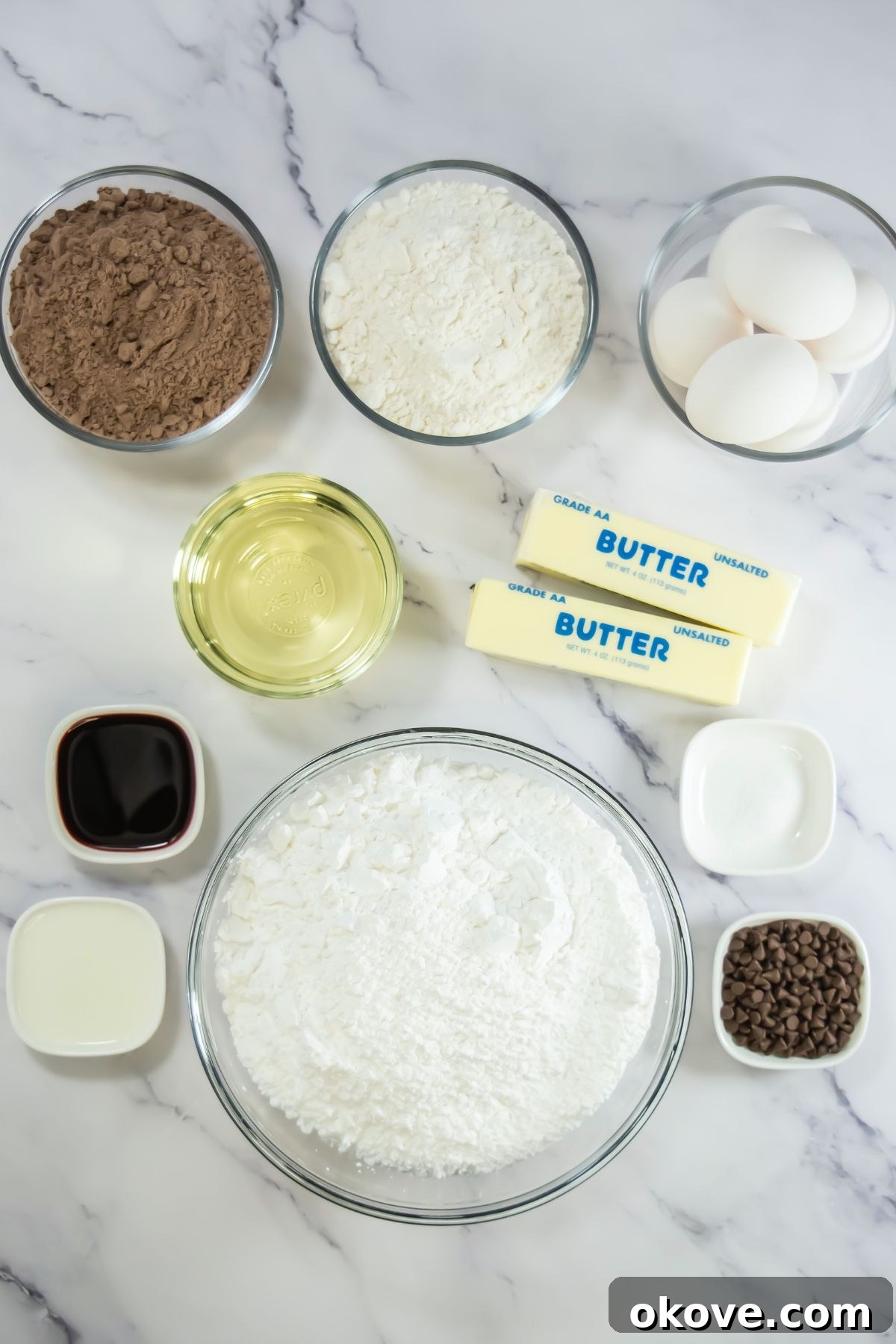 Neapolitan Swirl Cupcakes 3 All the necessary ingredients for baking Neapolitan cupcakes and making the strawberry frosting are neatly measured in small glass bowls, arranged on a pristine marble countertop, ready for preparation.