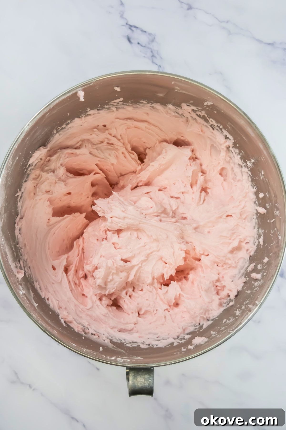 Neapolitan Swirl Cupcakes 7 A vibrant swirl of pink buttercream frosting fills a large silver mixing bowl, showcasing its smooth, creamy texture and inviting color, ready for decorating.