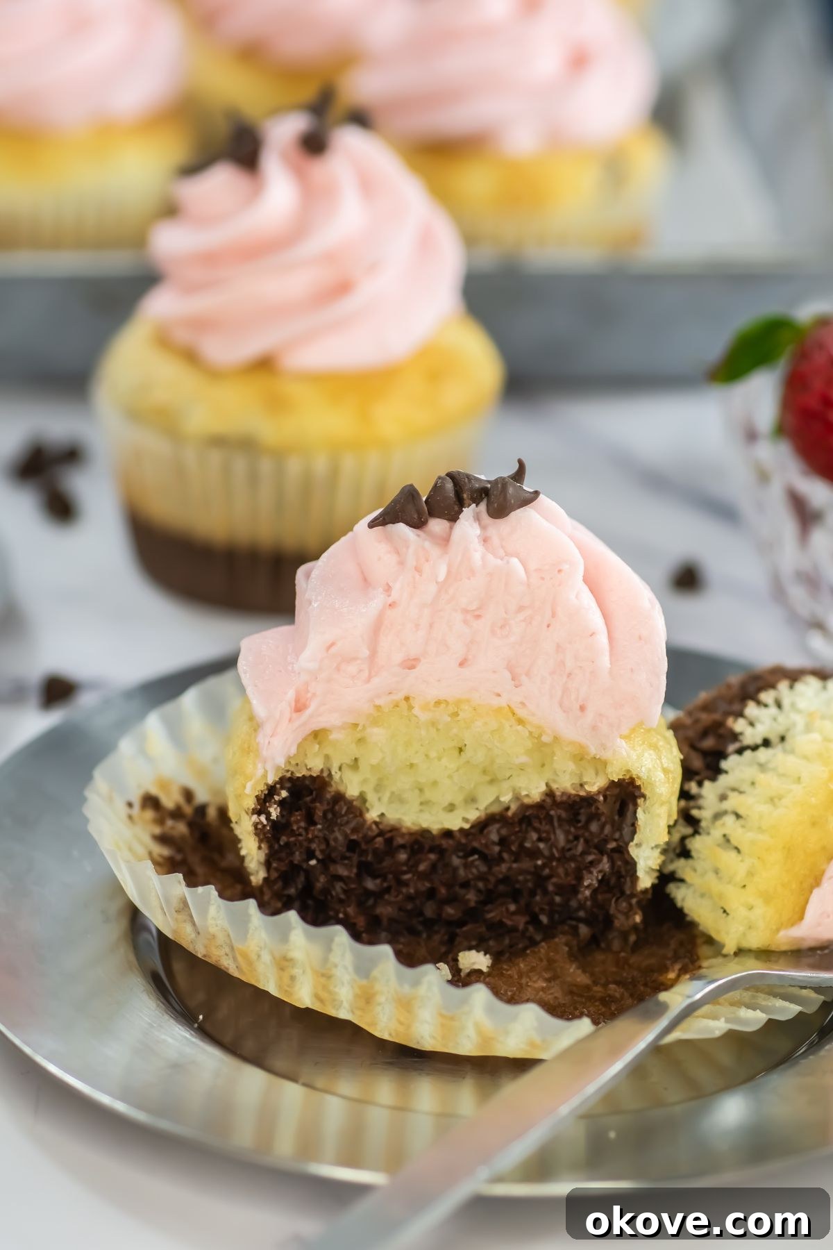 Neapolitan Swirl Cupcakes 9 A trio of perfectly layered Neapolitan cupcakes, featuring distinct chocolate and vanilla cake layers crowned with fluffy pink strawberry frosting, are artfully presented on a shimmering silver plate, ready to be enjoyed.