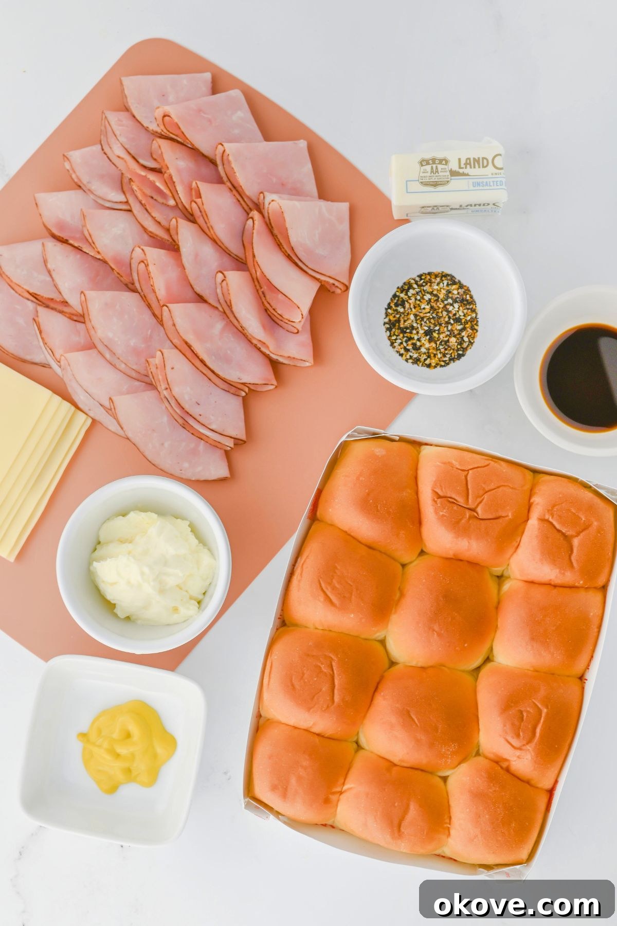 Array of fresh ingredients for ham and cheese sliders, including ham, Swiss cheese, mayonnaise, mustard, Hawaiian rolls, butter, Worcestershire sauce, and everything bagel seasoning, neatly arranged on a marble countertop.