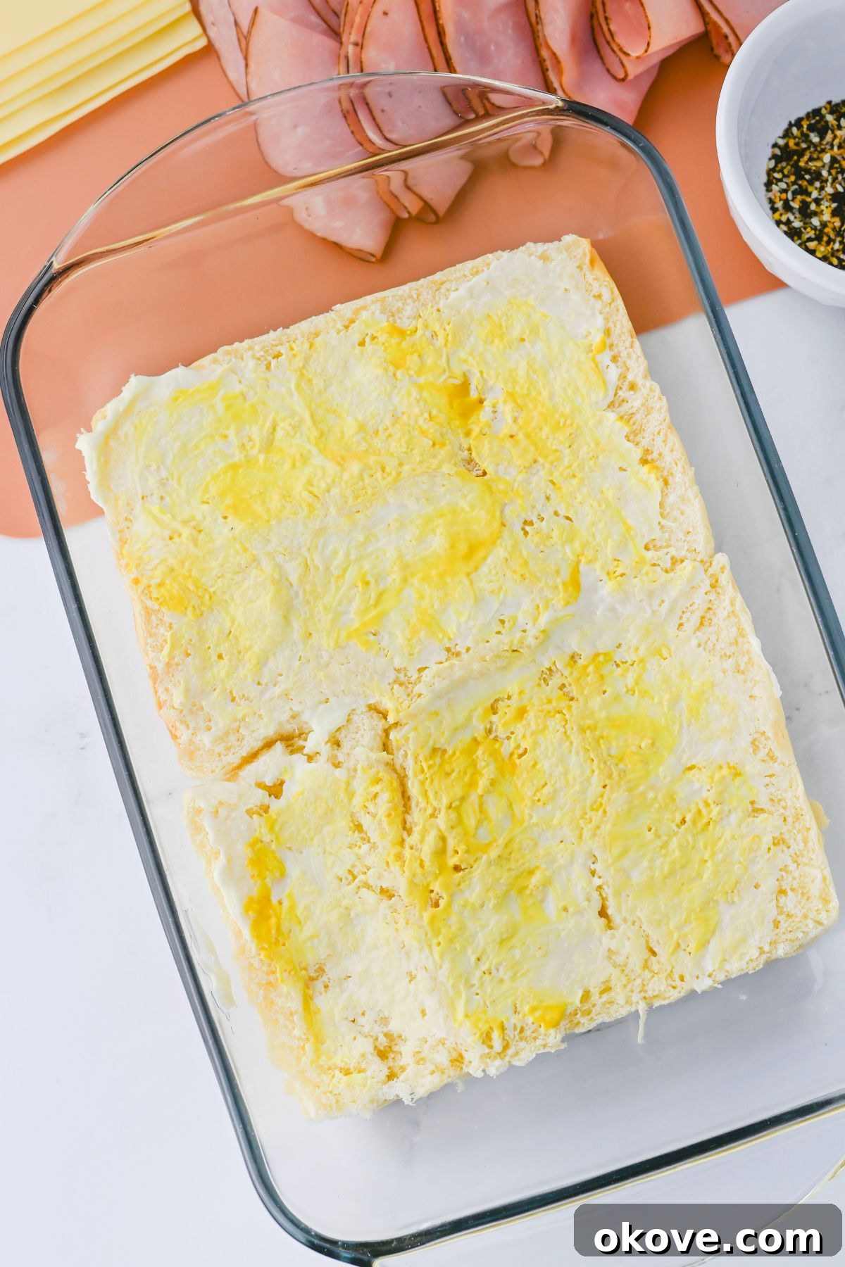 Bottom half of Hawaiian rolls spread with mayonnaise and mustard in a baking dish, ready for layering.
