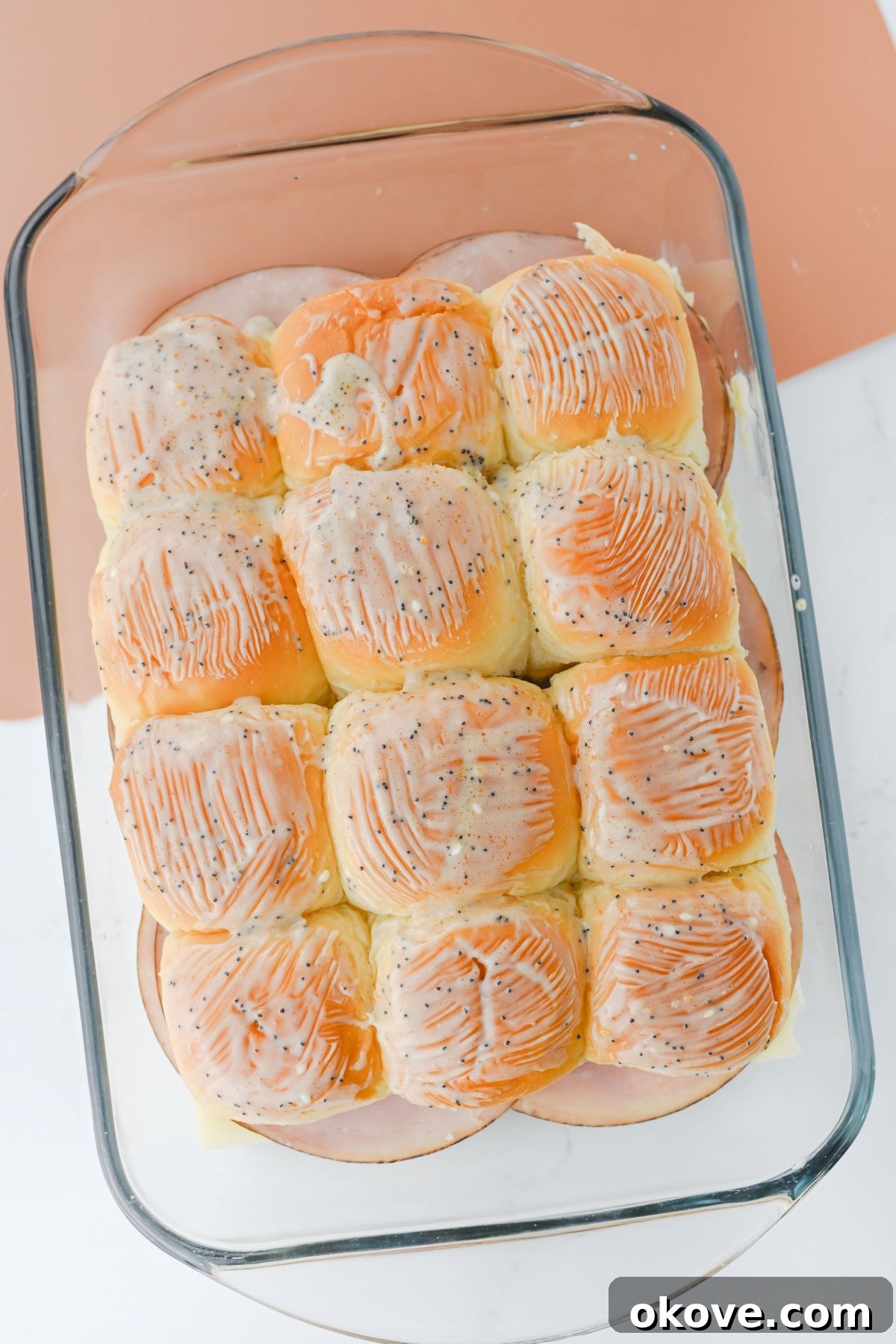 Melted butter glaze with seasonings being brushed over the tops of ham and cheese sliders in a baking dish.
