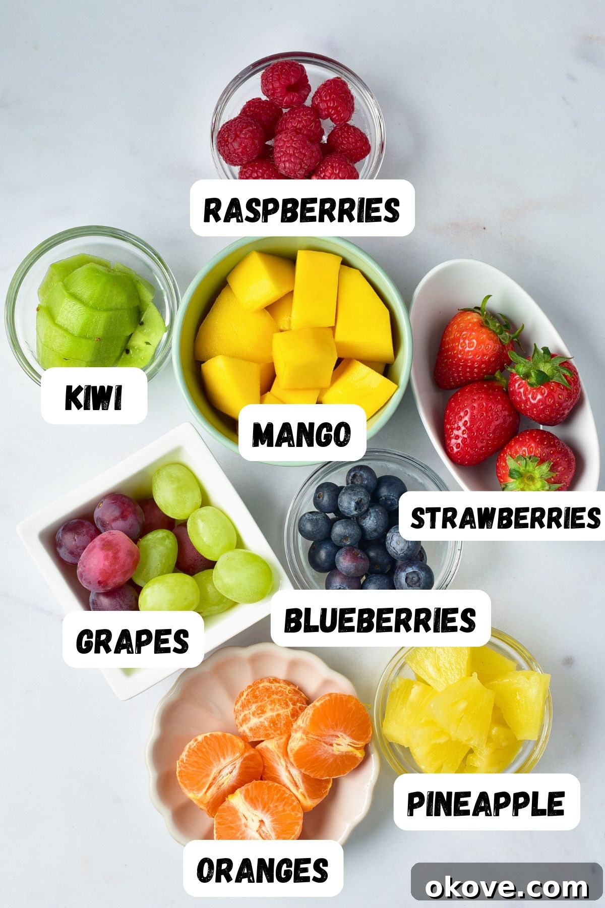 An assortment of fresh, brightly colored fruits, neatly cut and placed in small white bowls on a pristine marble countertop, ready for assembly.