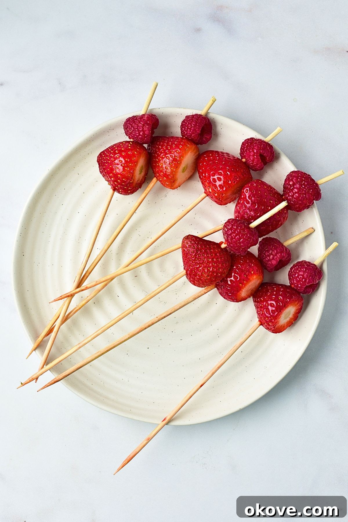 A wooden skewer with a raspberry and a strawberry, carefully threaded, resting on a white plate, showcasing the initial steps of assembly.