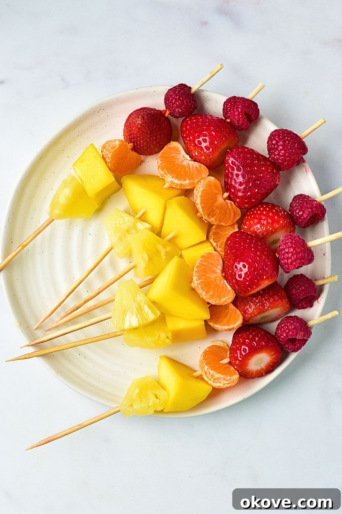 A wooden skewer with a colorful sequence of fruit slices, including mandarin, mango, pineapple, kiwi, green grape, and pink grape, laid out on a white plate, ready for the final touch.