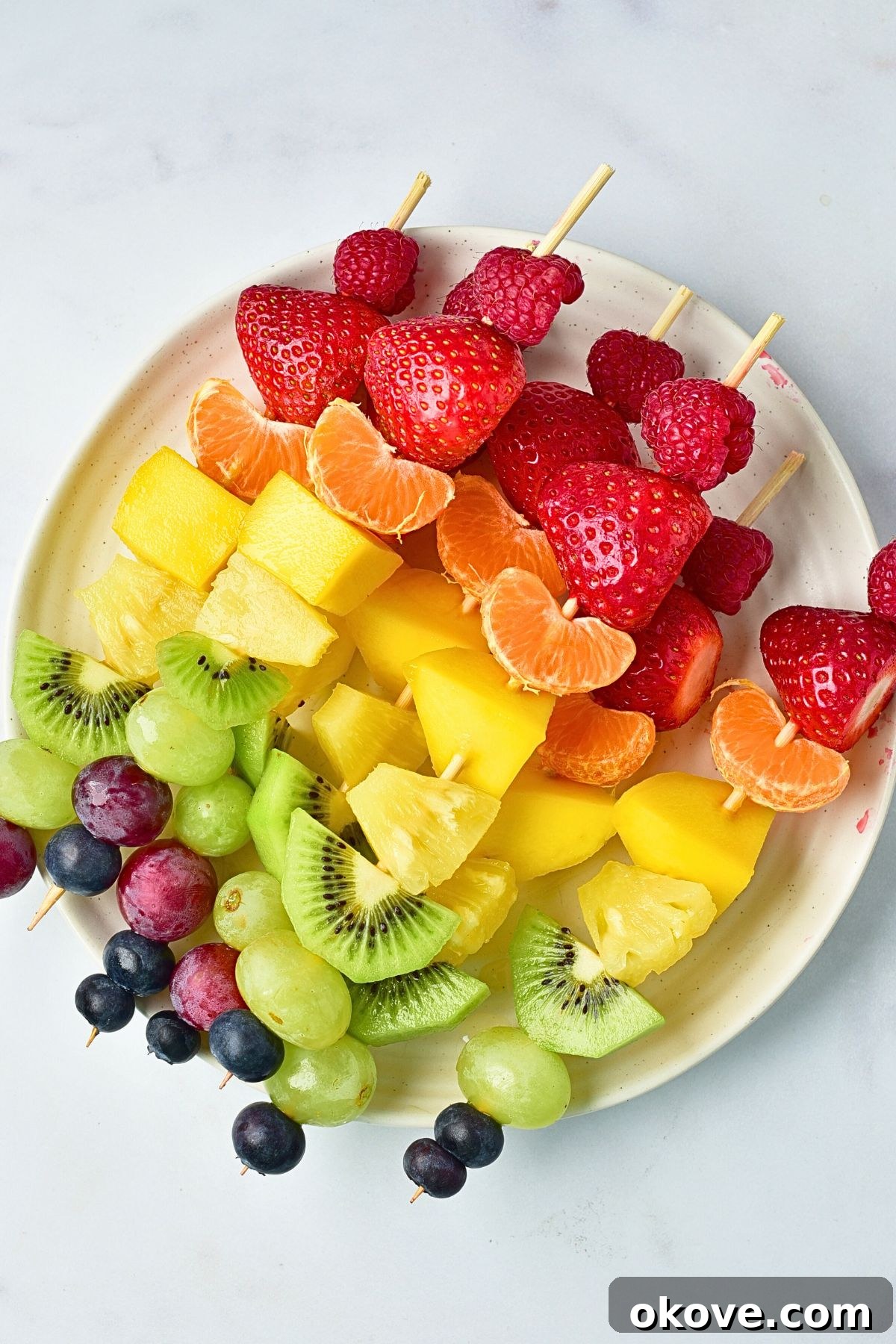 A completed fruit skewer, topped with a blueberry, stands upright among other colorful skewers on a white plate, showcasing the vibrant final product.