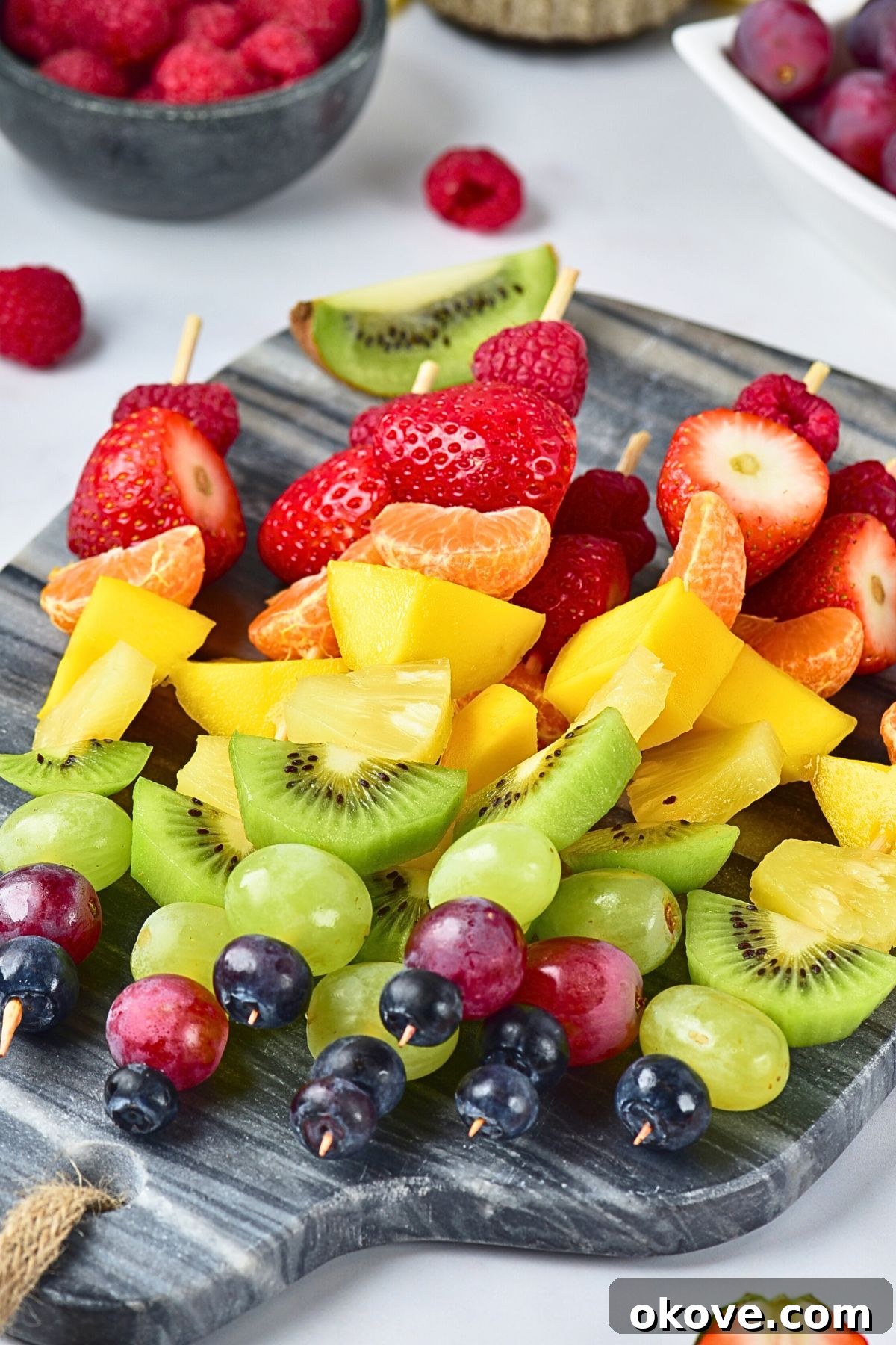 Several finished fruit skewers, each meticulously threaded with a rainbow of colors, arranged artfully on a small light gray cutting board, ready to be served.