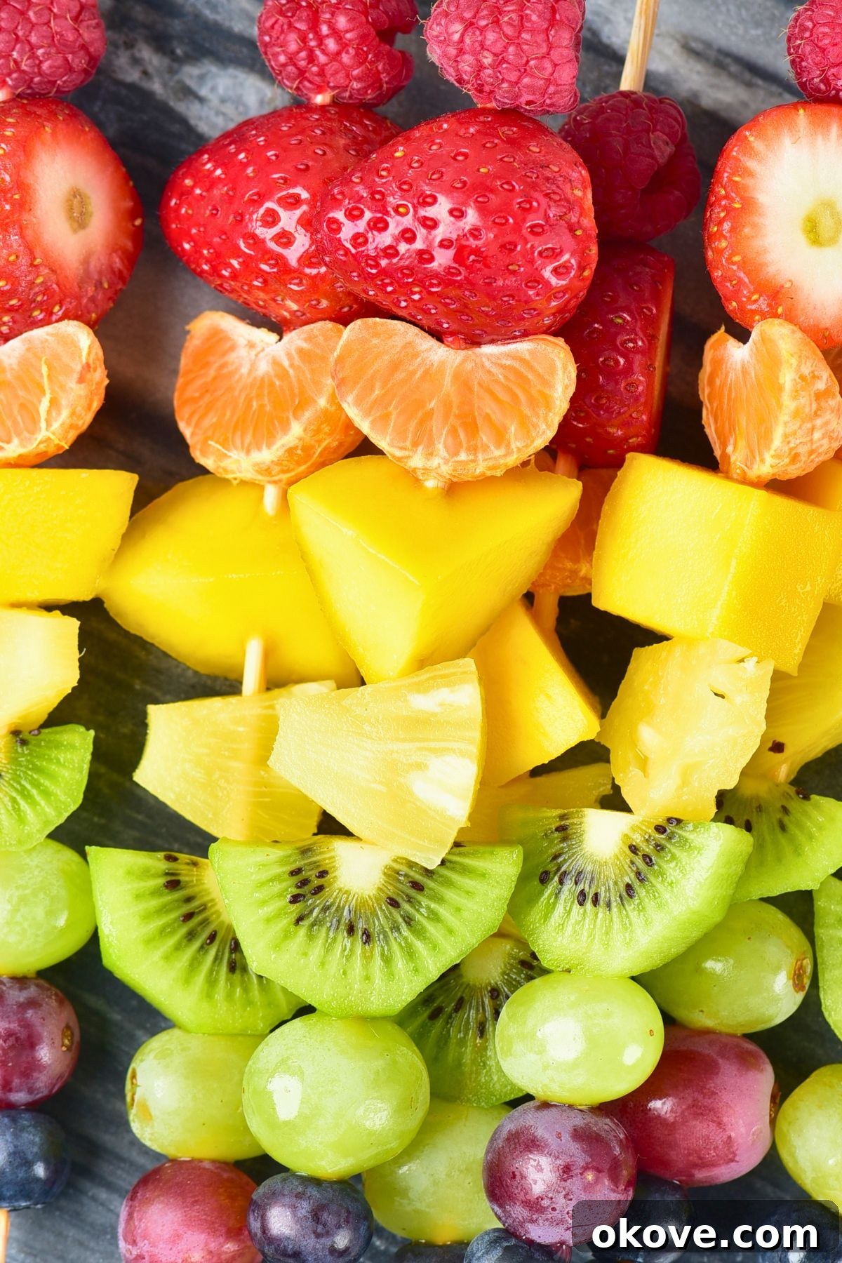 A close-up shot of multiple vibrant fruit skewers, arranged meticulously in a rainbow pattern, resting on a small light gray cutting board, highlighting their freshness and appeal.