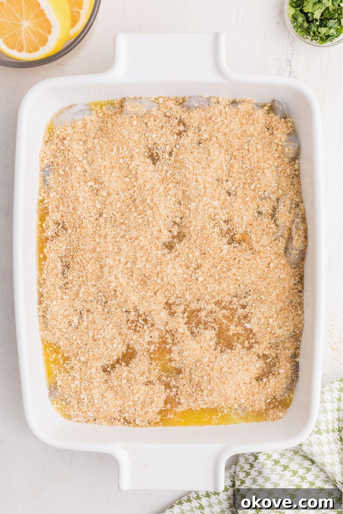 Shrimp in a white baking dish, generously topped with a rich melted butter and seasoning mixture, followed by a sprinkle of crispy breadcrumbs and Parmesan cheese.