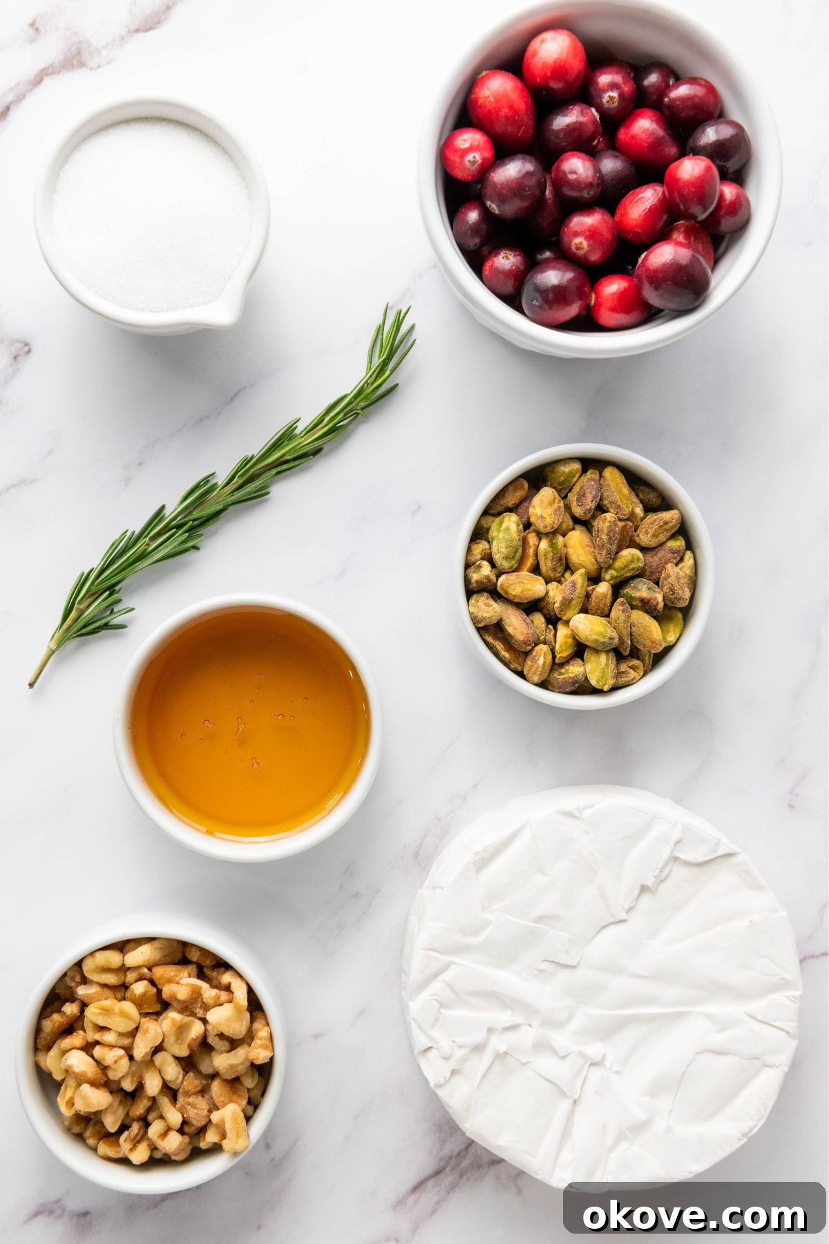 Warm Brie Delight 3 Carefully measured ingredients for baked brie, including a wheel of brie, a bowl of fresh cranberries, chopped walnuts, pistachios, a jar of golden honey, and fragrant rosemary sprigs, all arranged on a white marble surface.