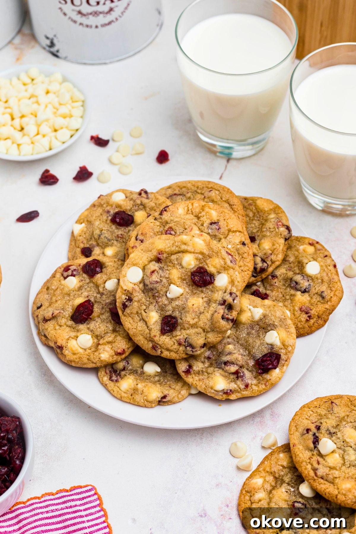 Stacked white chocolate chip and cranberry cookies on a white plate with glasses of milk in the background, invitingly presented.