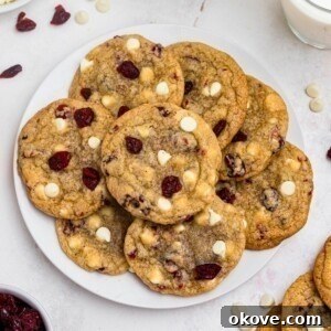 White chocolate chip and cranberry cookies stacked on a white plate with glasses of milk on the table, ready to be enjoyed.