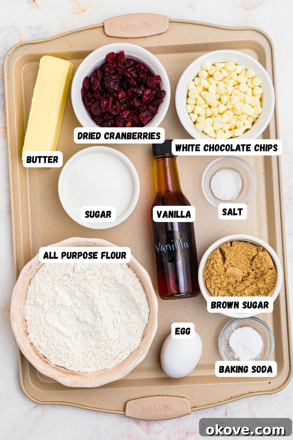 Ingredients for white chocolate cranberry cookies, precisely measured into small bowls and arranged on a baking sheet on a sleek marble table, awaiting preparation.