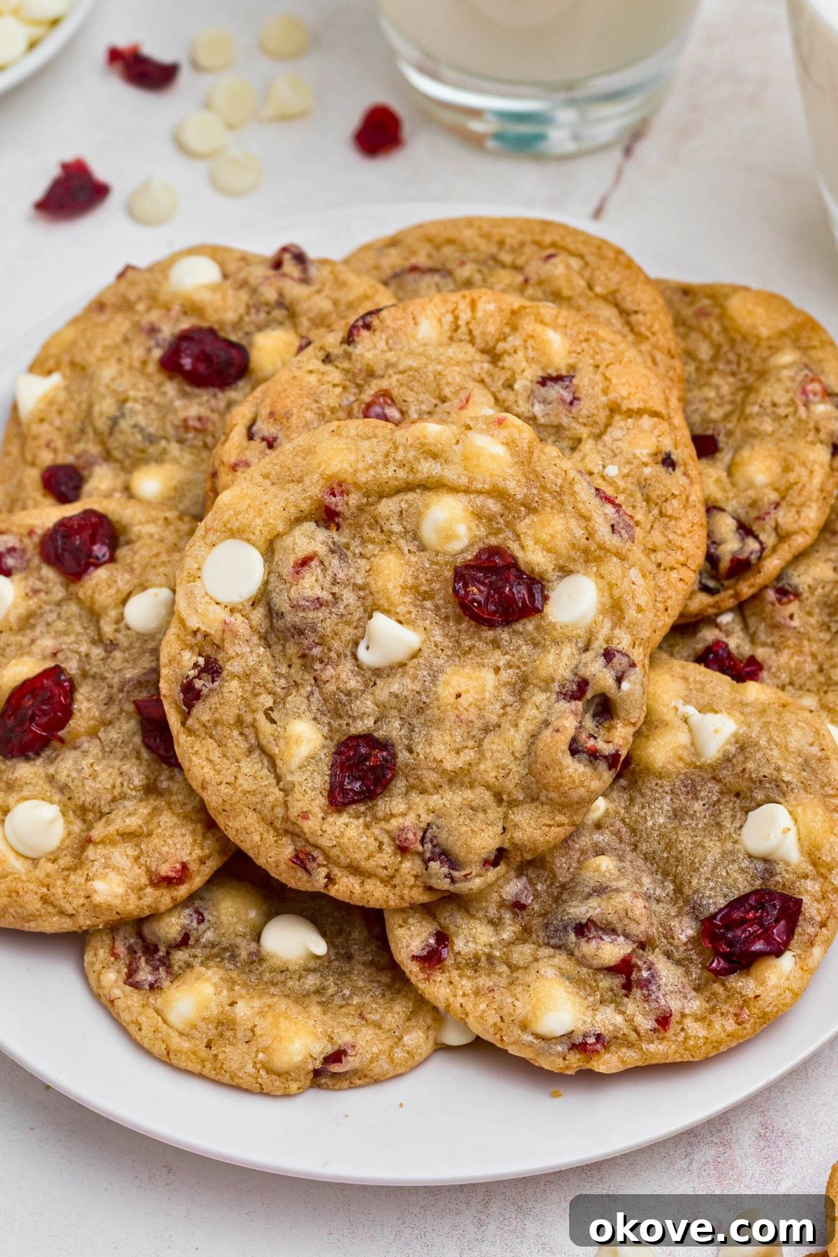 A beautifully arranged stack of golden white chocolate chip and cranberry cookies on a pristine white plate, with blurred glasses of milk in the background, signaling a perfect treat.