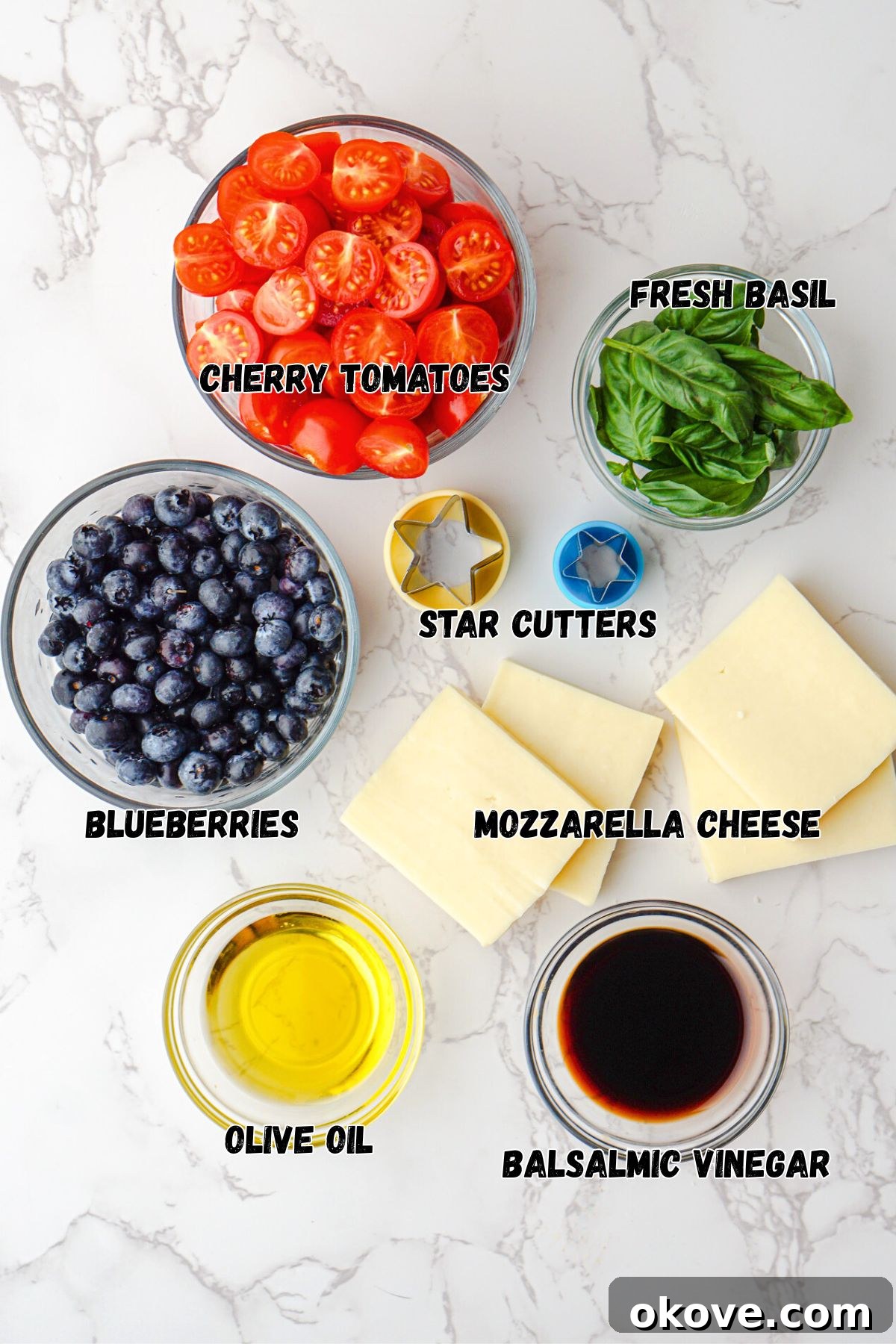 Individual fresh ingredients required to make Berry Caprese Salad, including halved cherry tomatoes, plump blueberries, a block of fresh mozzarella, and a bunch of green basil leaves, presented in small glass bowls on a white marble tabletop.