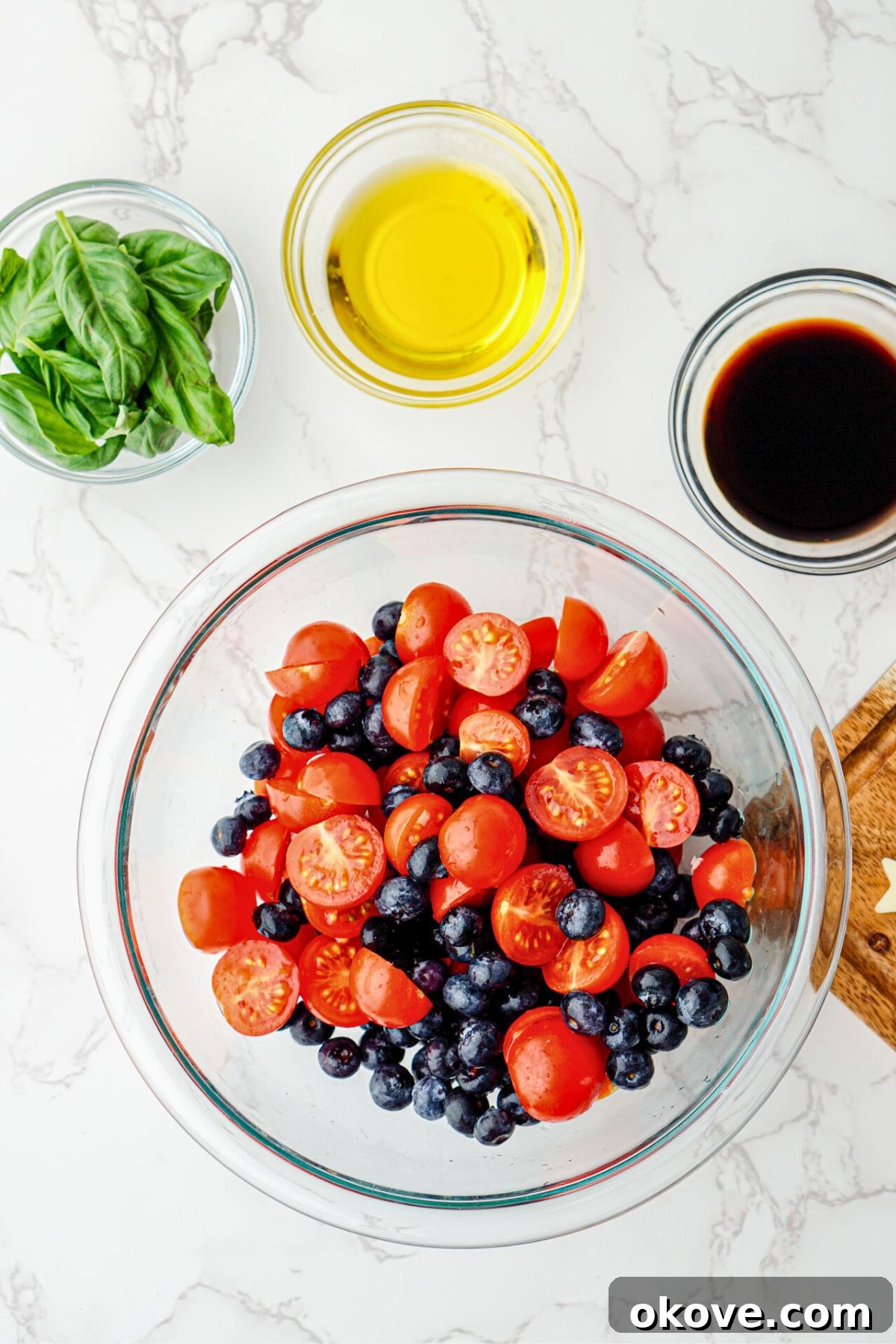 Halved cherry tomatoes and whole blueberries being gently mixed together in a clear glass serving dish, forming the red and blue base of the salad.