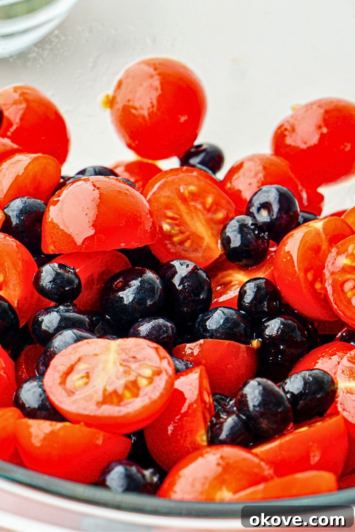 Cherry tomatoes and blueberries in a clear glass bowl, freshly tossed with torn basil leaves, olive oil, and balsamic vinegar, ready for the next step.
