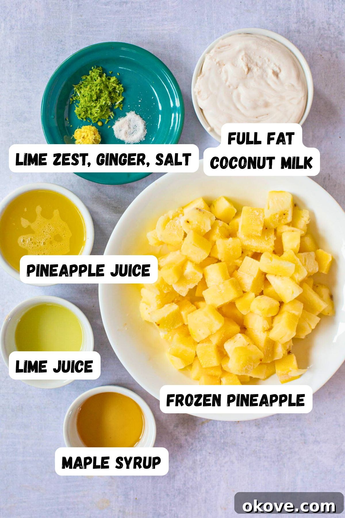Ingredients needed to make virgin pina colada measured in small white bowls on a marble table, including frozen pineapple, coconut milk, pineapple juice, lime, ginger, and maple syrup.