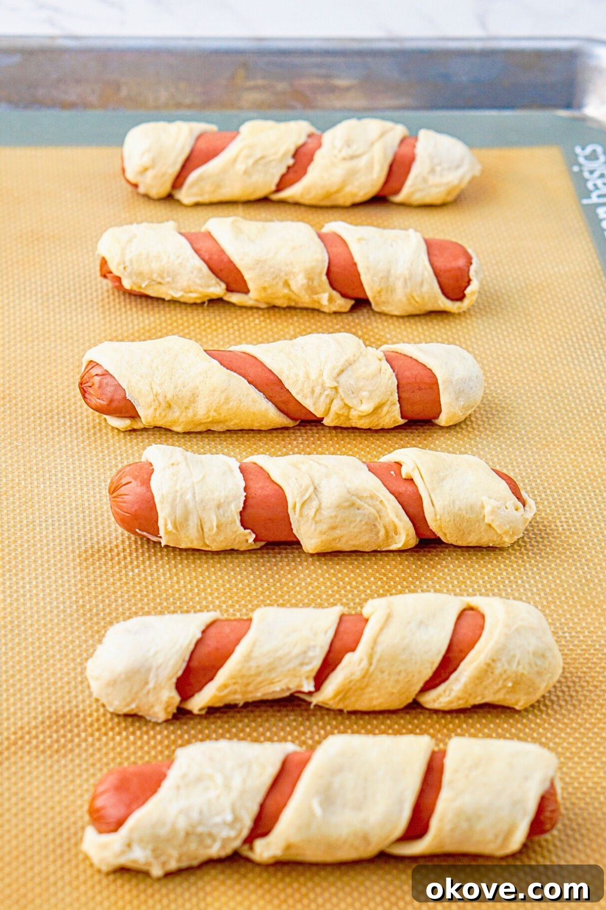 Hot dogs wrapped in strips of crescent roll dough before baking.